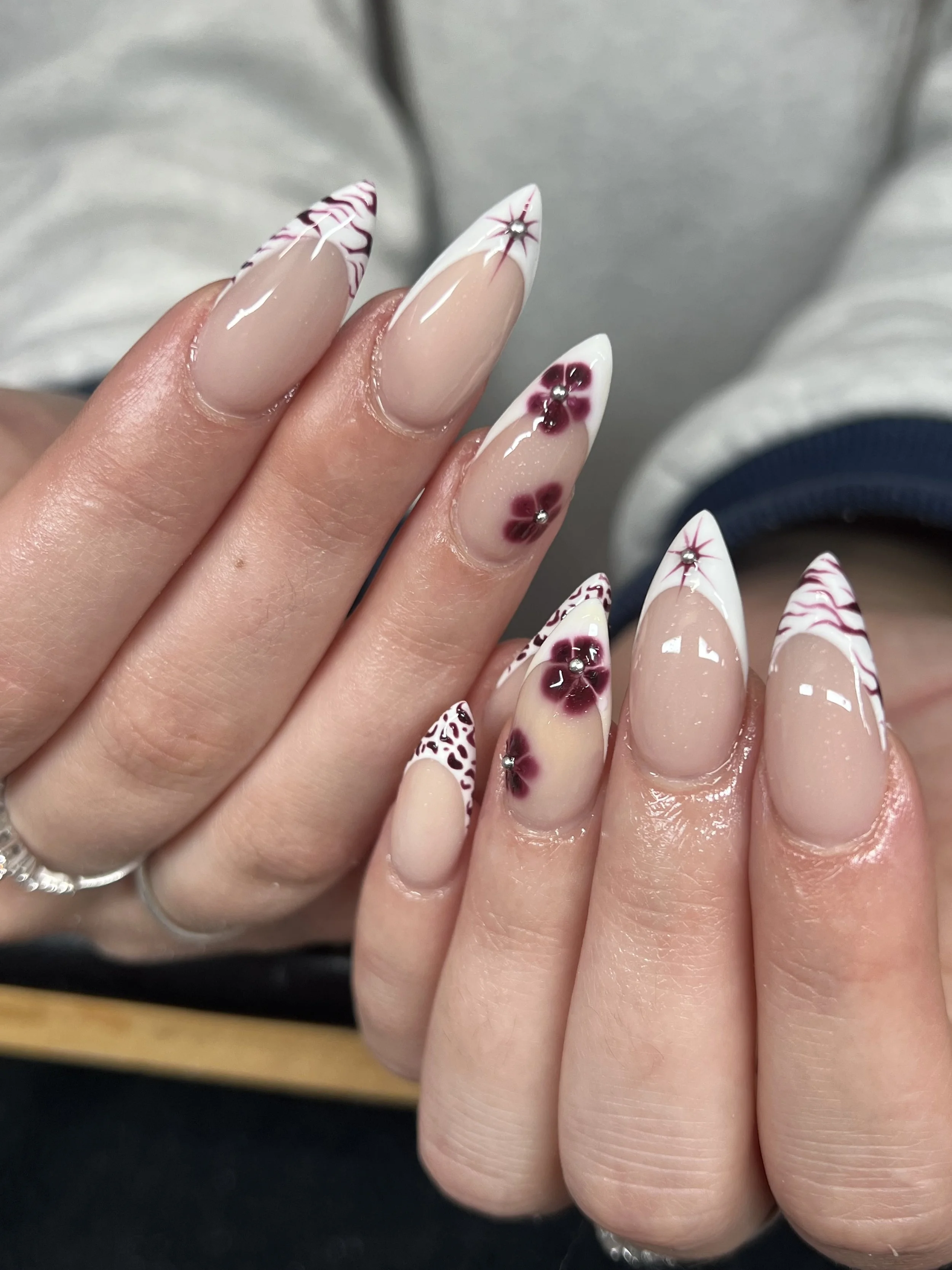 Close-up of a person's hands with intricate, decorated nail art featuring a nude base, white tips with red and black patterns, and floral designs with rhinestone accents.