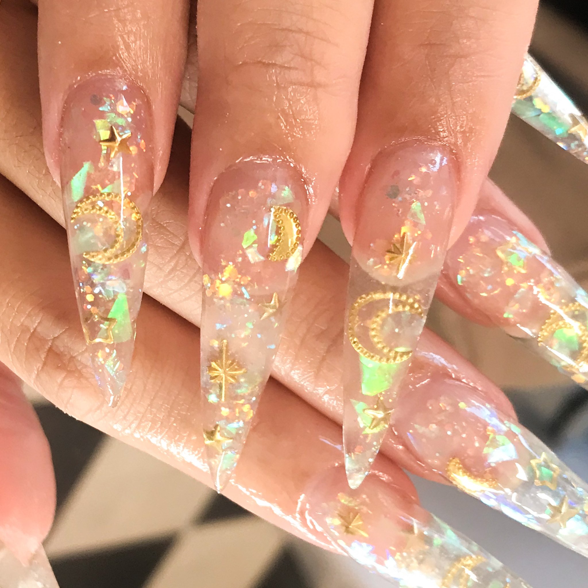 Close-up of long, clear acrylic nails decorated with gold star, moon, and crescent moon shapes, along with iridescent holographic flakes.