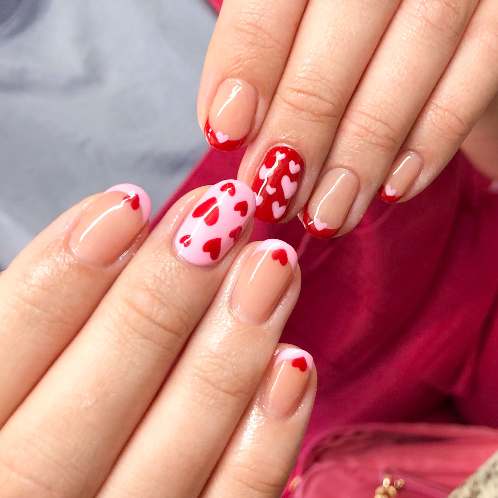 Close-up of manicured fingernails with Valentine’s Day themed nail art, featuring red, pink, and nude colors with heart designs.
