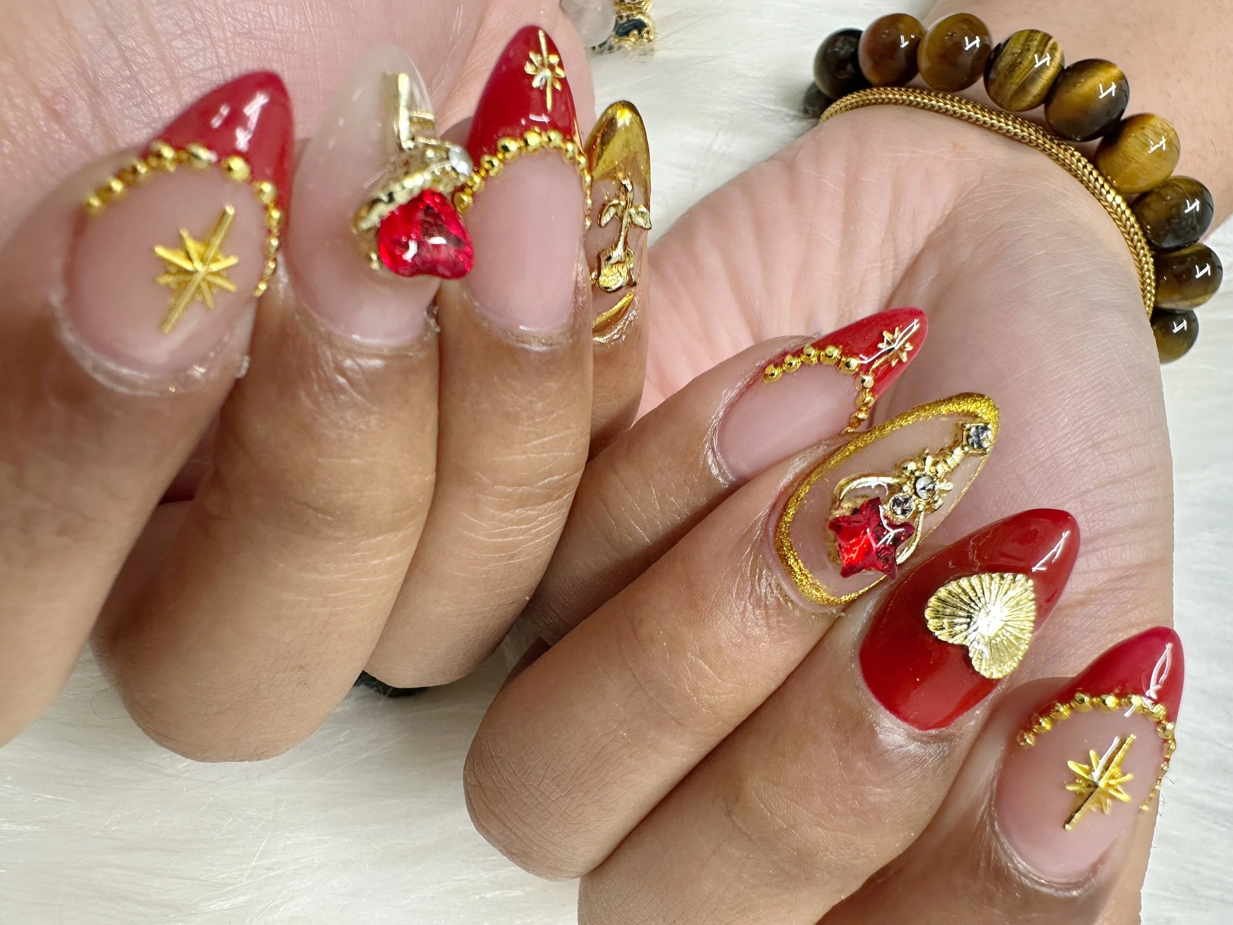 Close-up of decorated fingernails with red, gold, and clear polish, featuring gold accents, charms, and embellishments, along with gold rings and wooden bead bracelet.