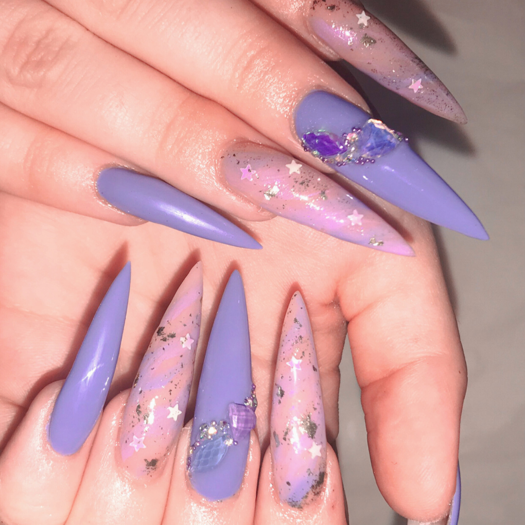 Close-up of long, stiletto-shaped acrylic nails with lavender polish, decorated with star-shaped and glitter accents, and some nails featuring 3D embellishments like purple hearts and pearls.
