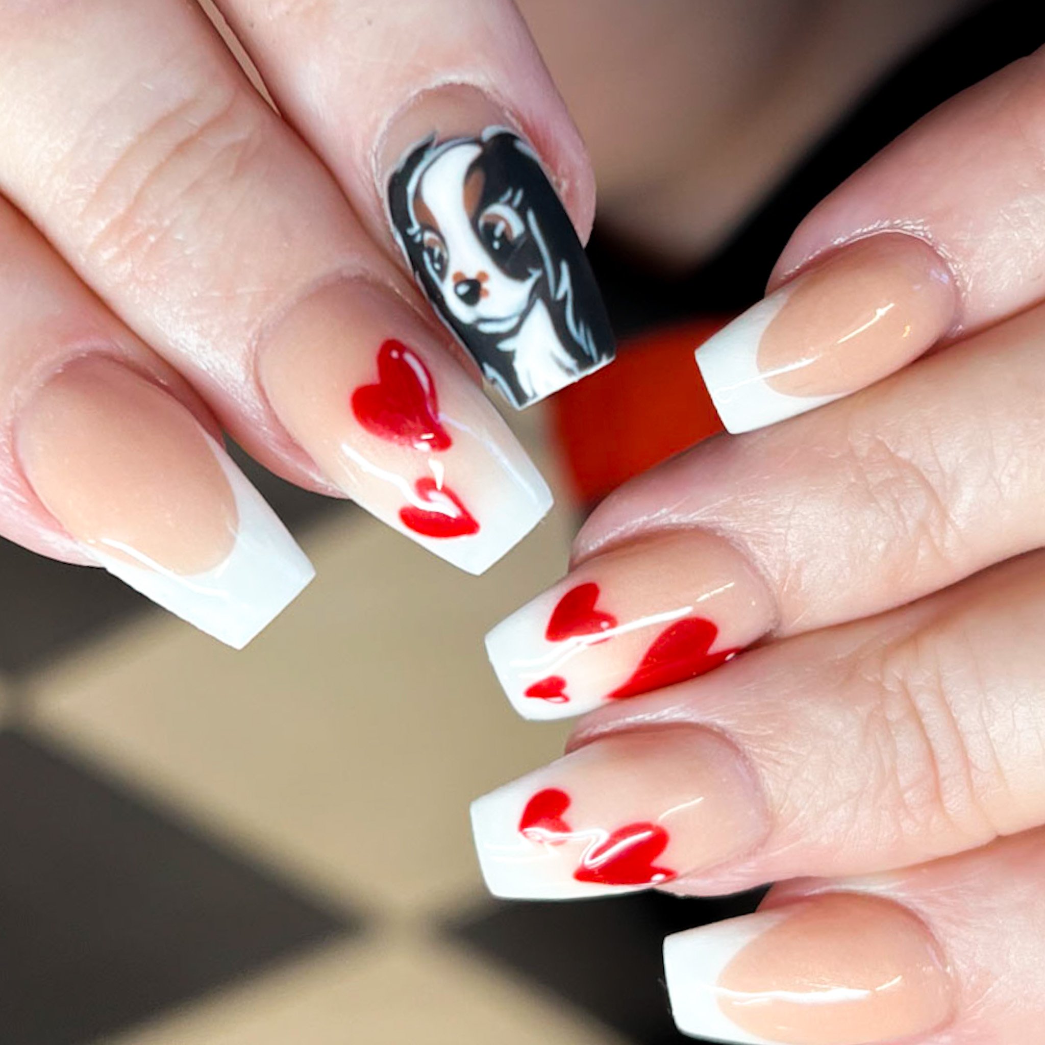 Nail art with red hearts on a clear and French tip background, and a black and white dog painted on the thumbnail.