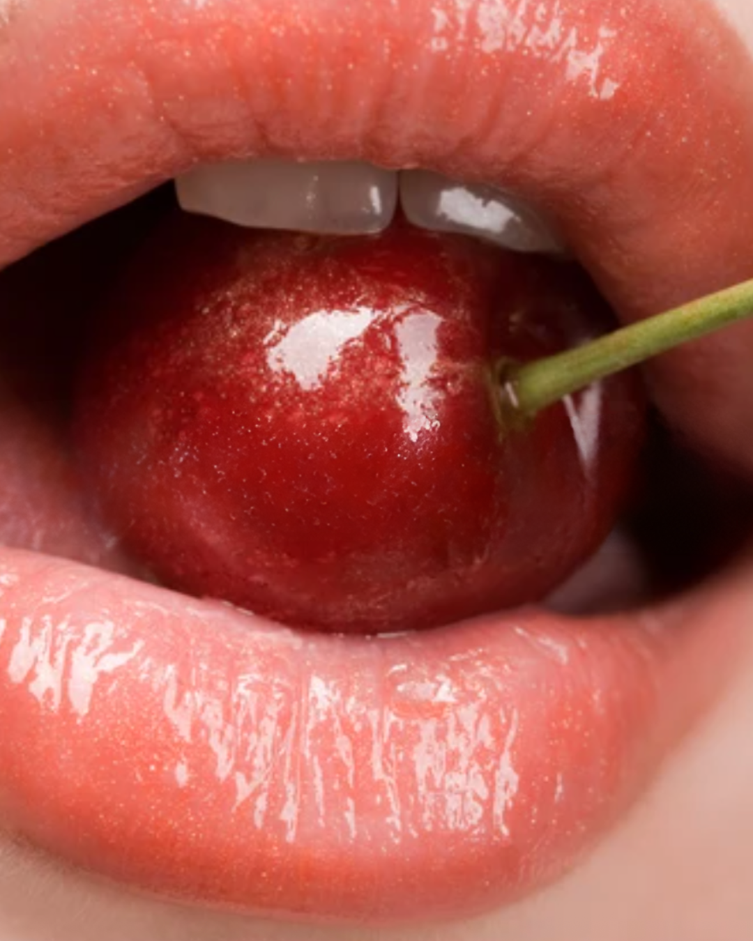 Close-up of lips with shiny pink lipstick, a cherry held between the lips with a green stem, and the tongue touching the cherry.