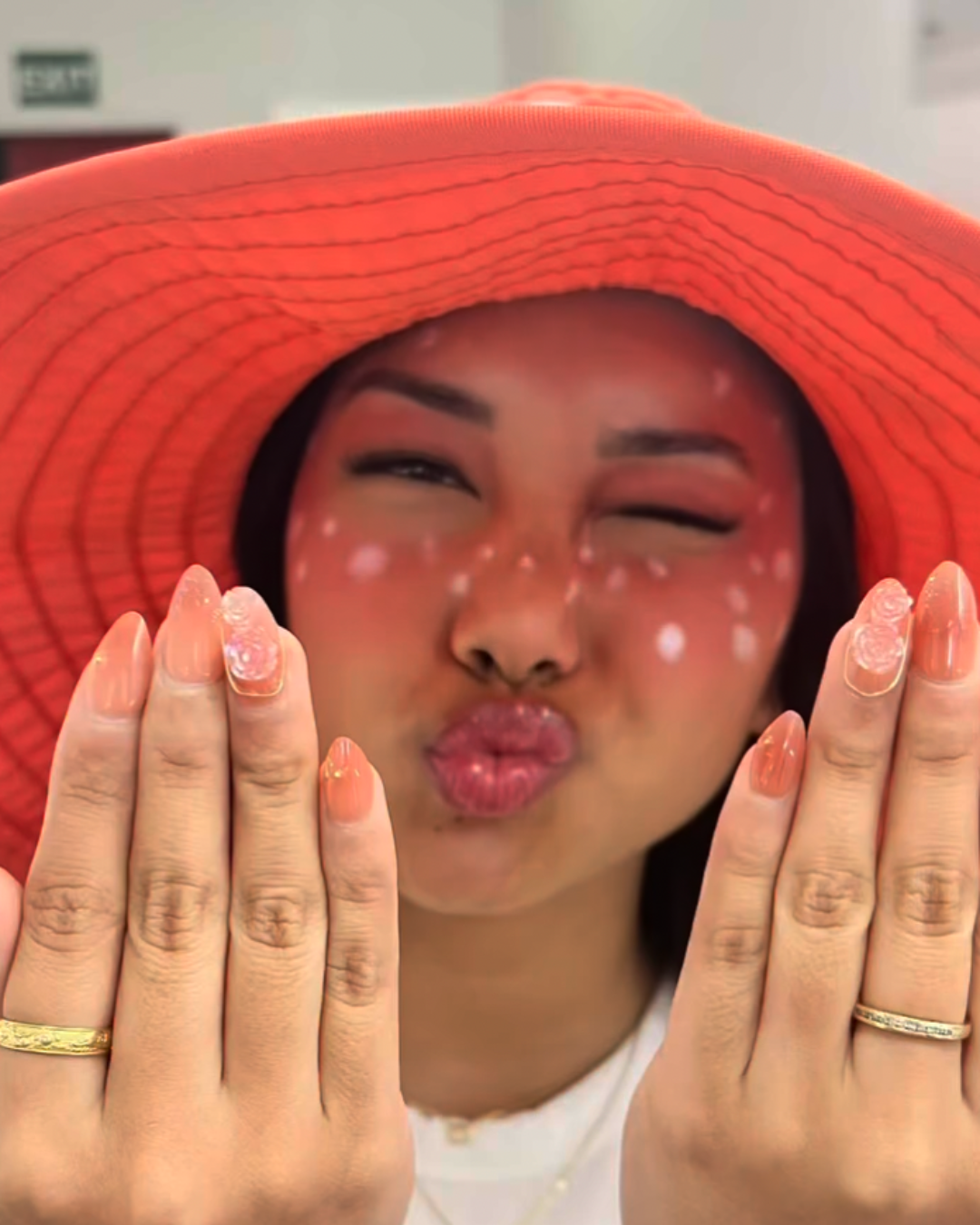 A woman wearing a large red sun hat, making a kissy face while holding out her hands with pink and clear fingernails, with white spots on her face, in an indoor setting.