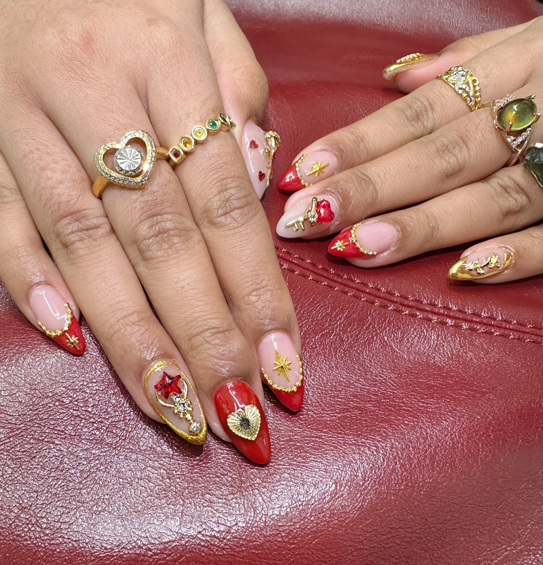 Hands with decorated nails featuring red, white, and gold nail art and multiple rings on fingers, resting on a red leather surface.