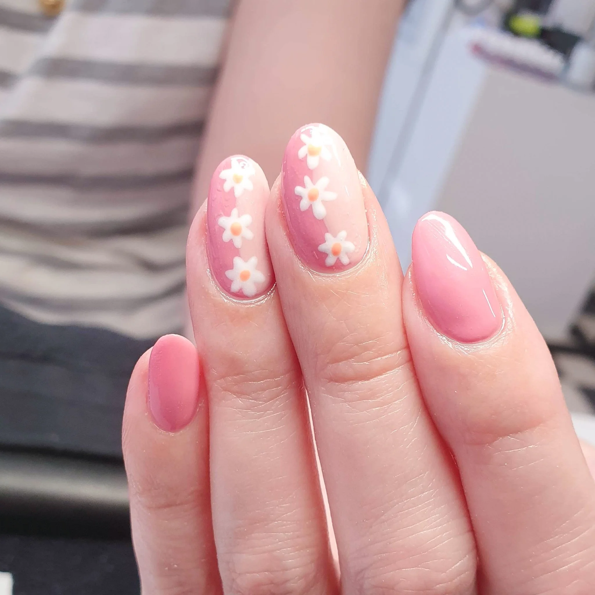 Close-up of a person's hand showing four fingers with manicured nails, featuring pink and nude polish with white daisy floral designs on the ring finger.