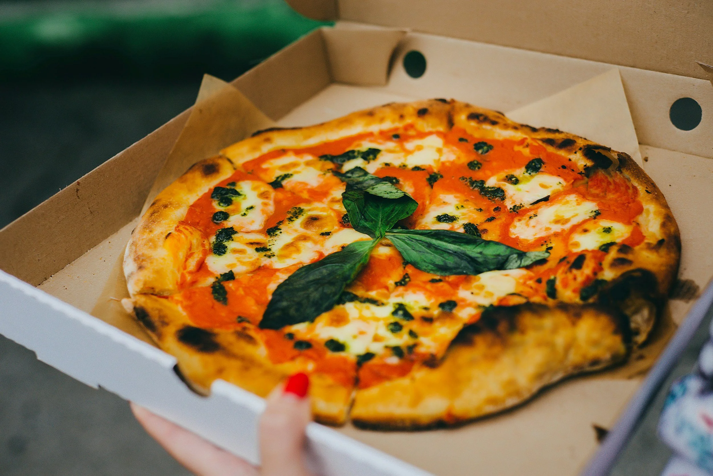 A pizza with mozzarella cheese, tomato sauce, basil leaves, and pesto on top, inside a pizza box.