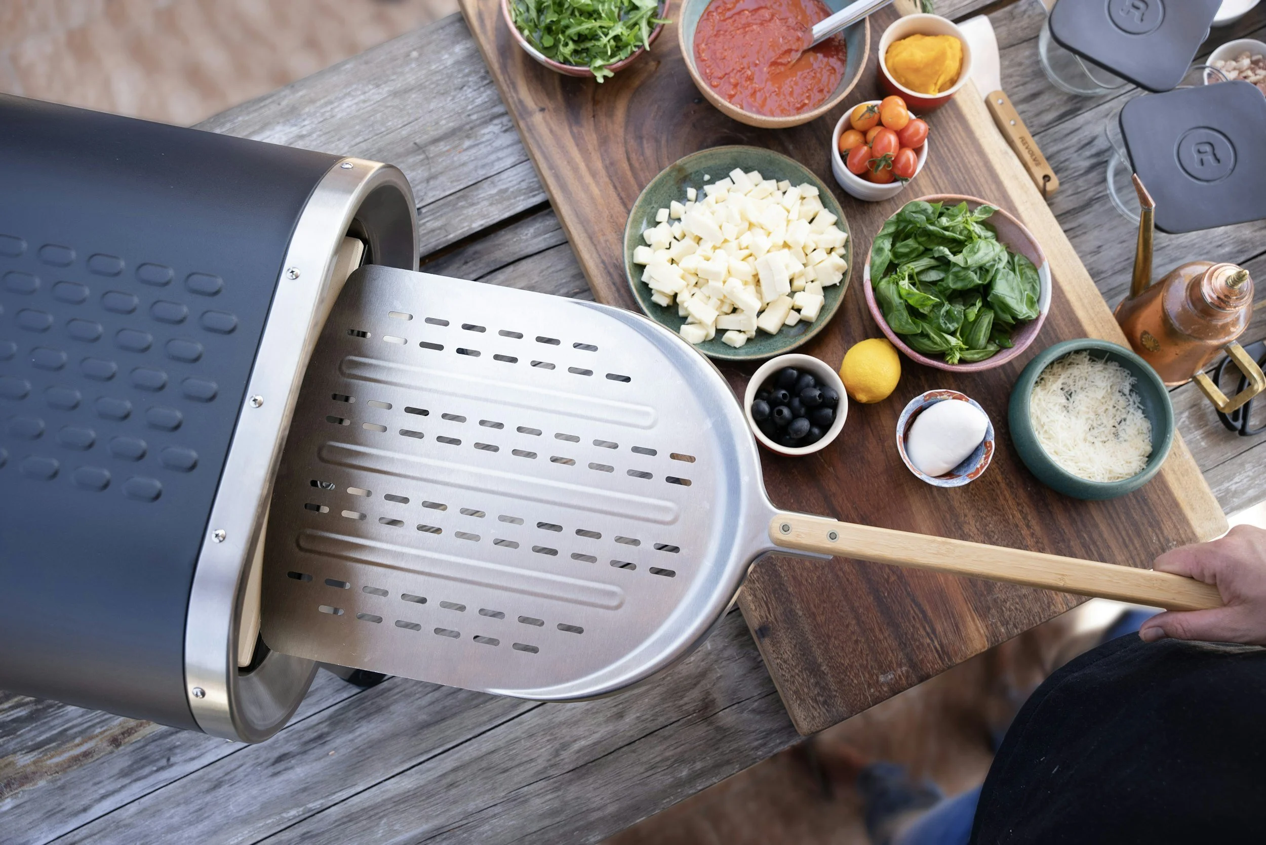 Grill with various bowls of ingredients for cooking, including cheese, cherry tomatoes, spinach, black olives, and sauces, on a wooden table.