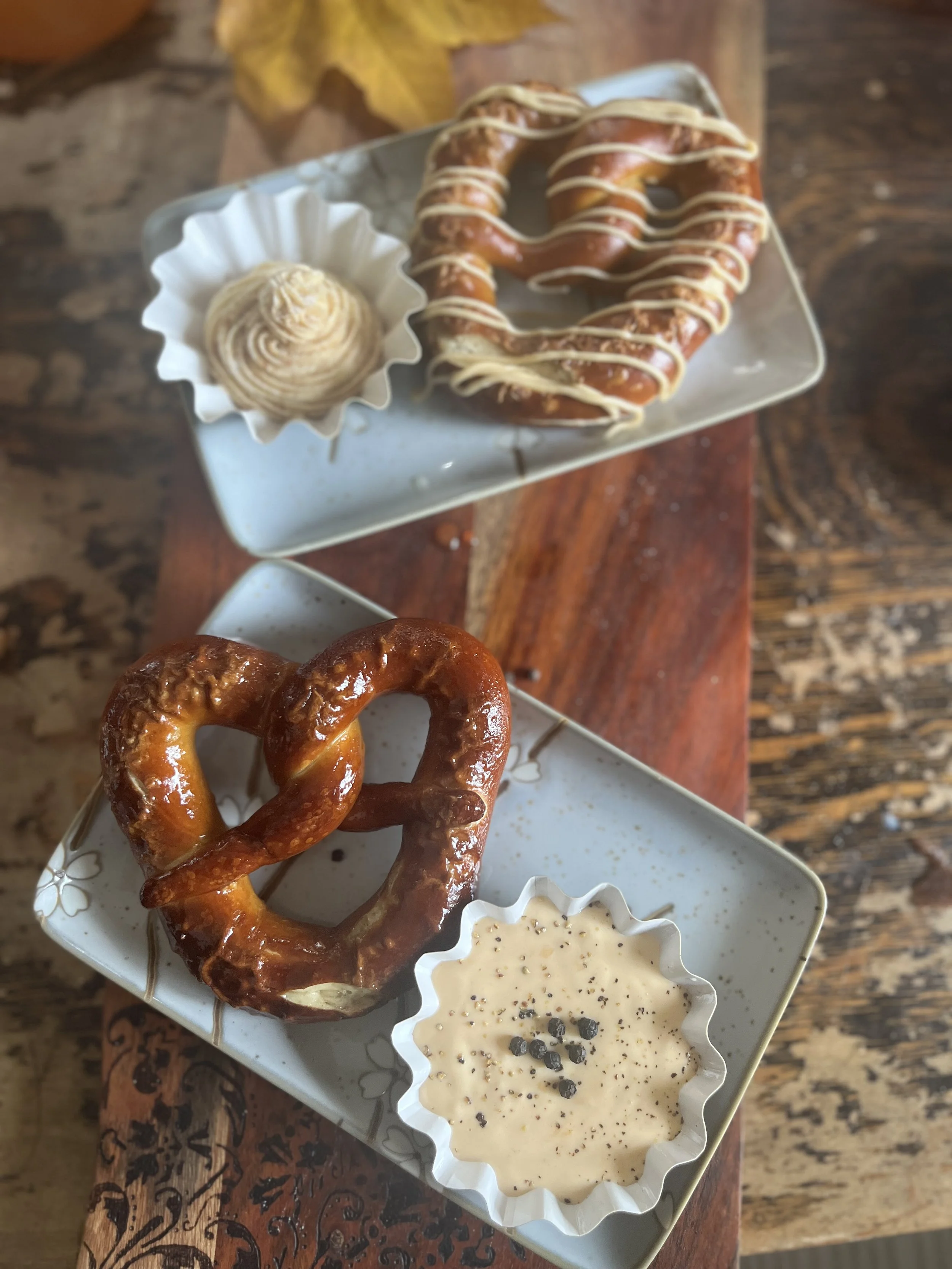 Two plates with pretzels, one with a pretzel with white icing and the other with a plain pretzel, accompanied by dipping sauces and a cinnamon roll on a wooden table.