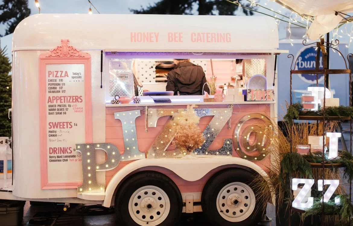 Pink food truck with a sign that reads "Honey Bee Catering" and a menu offering pizza, appetizers, sweets, and drinks. The truck has large illuminated letters spelling "JA" on the side with a flower decoration, and a side display shelf with decorative items and large letter lights spelling "ZZ".