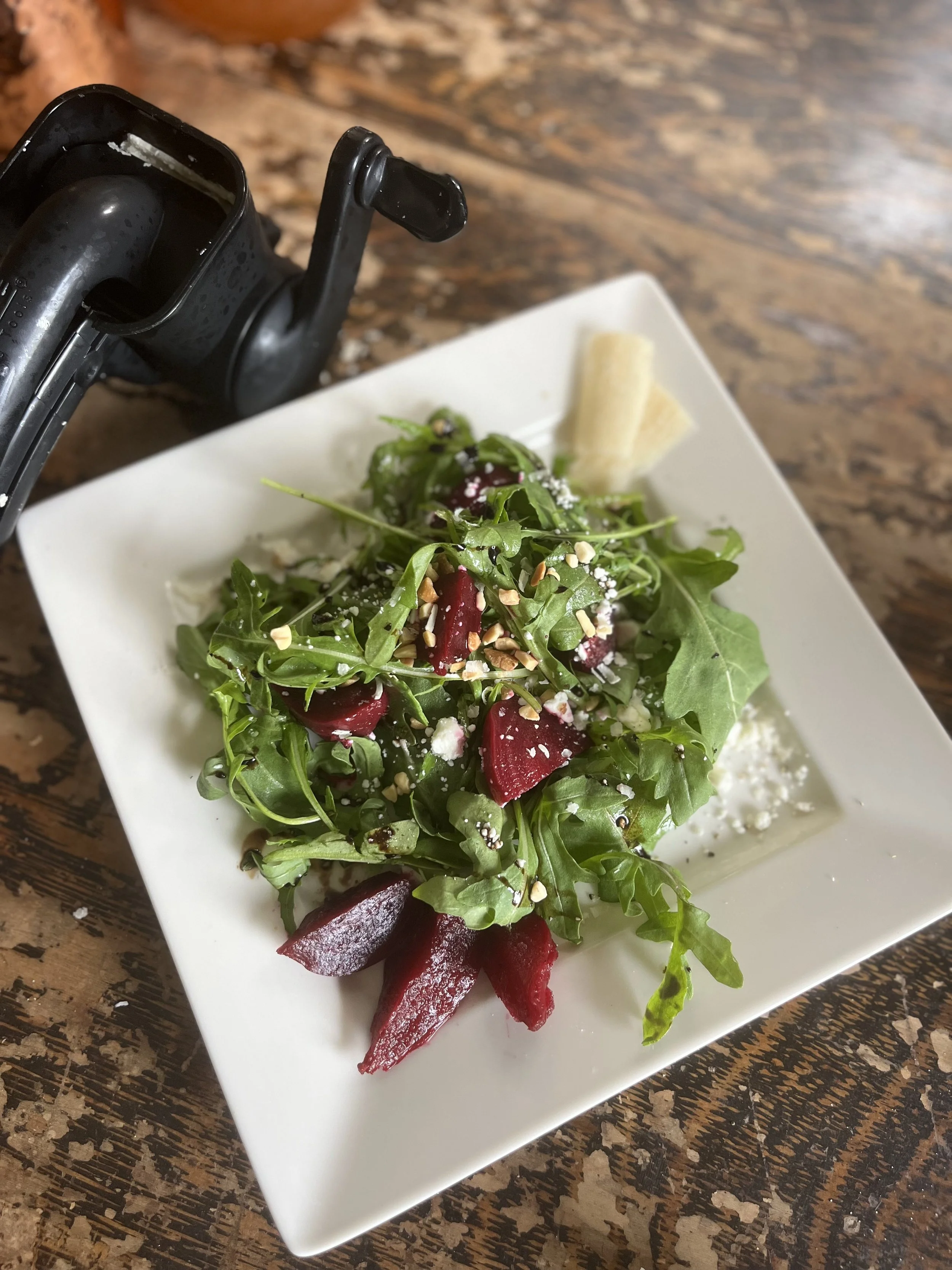 Mixed green salad with beets, nuts, grated cheese, and leafy greens on a white square plate, with a small piece of bread and a black camera lens nearby on a wooden table.