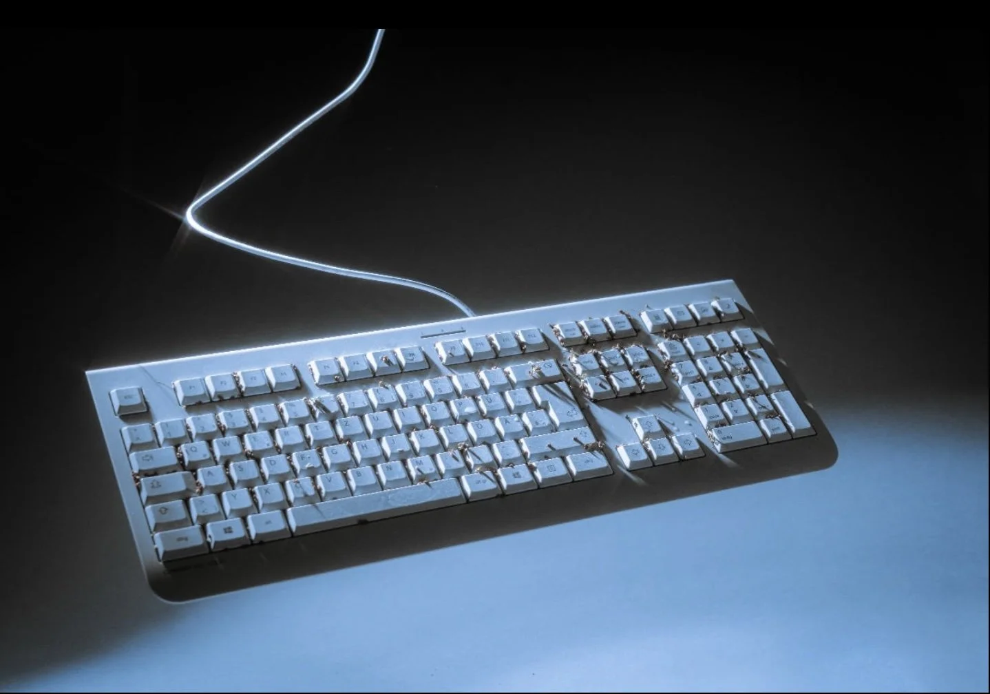 A damaged computer keyboard with a bent key and broken parts, connected to a power source with a nearby illuminated wire on a dark surface.
