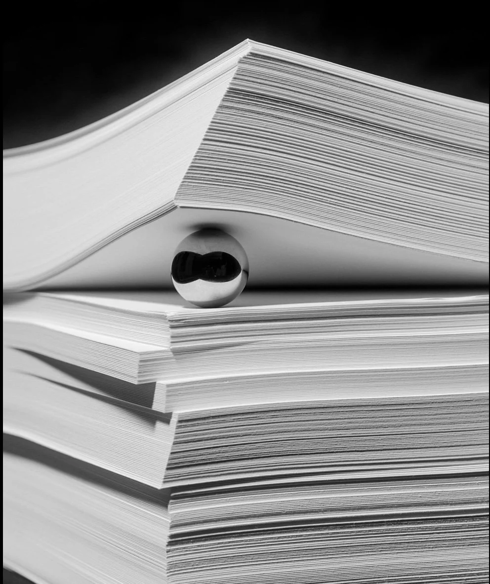 Black and white photo of a large open book resting on a stack of closed books with a reflective marble balancing on the open book's edge.