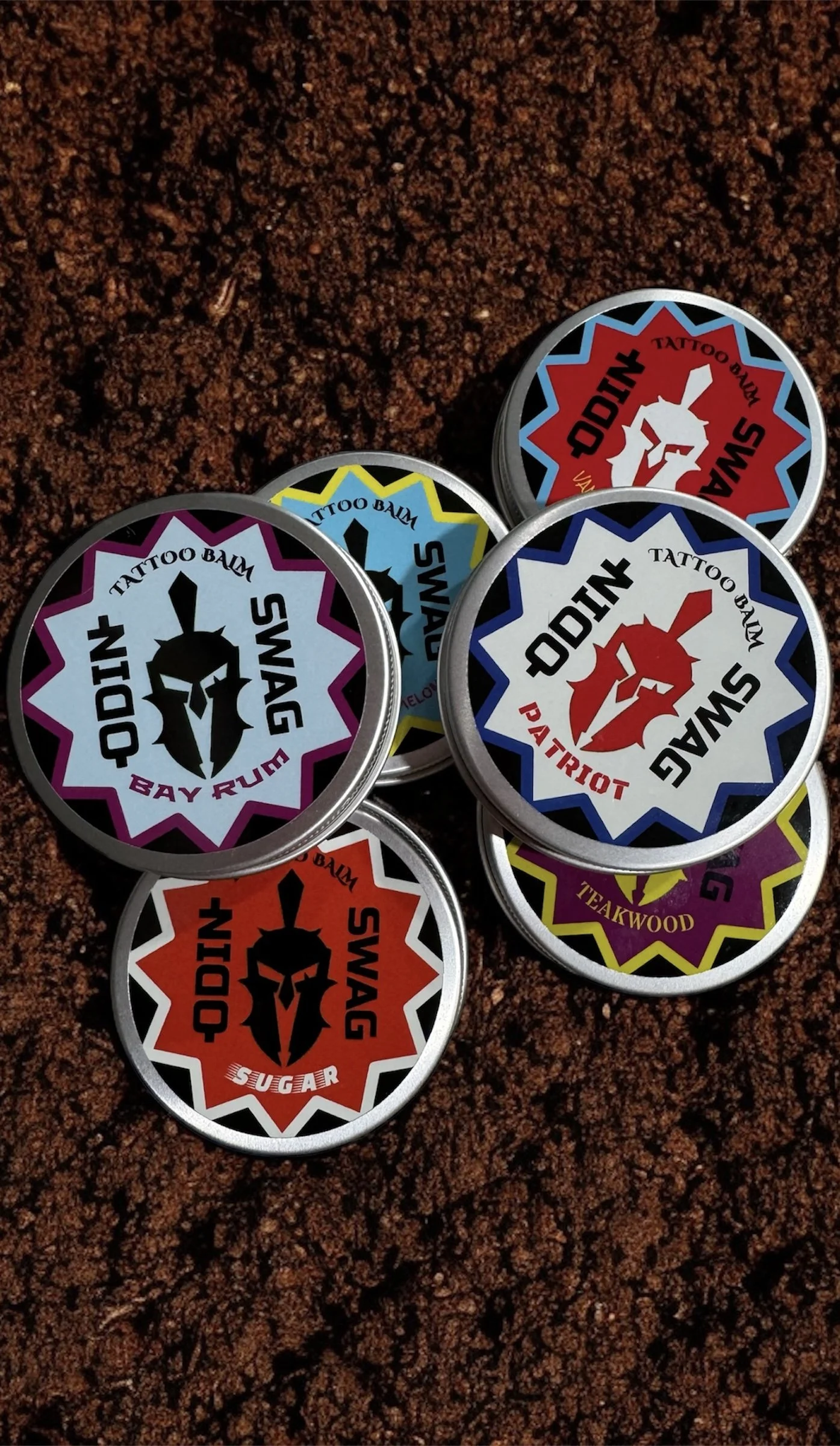 Five tins of Odin tattoo balm with colorful labels, featuring a Spartan helmet logo and names like 'Bay Rum,' 'Swag,' 'SUGAR,' 'Patriot,' and 'Takwod,' placed on reddish-brown textured ground.