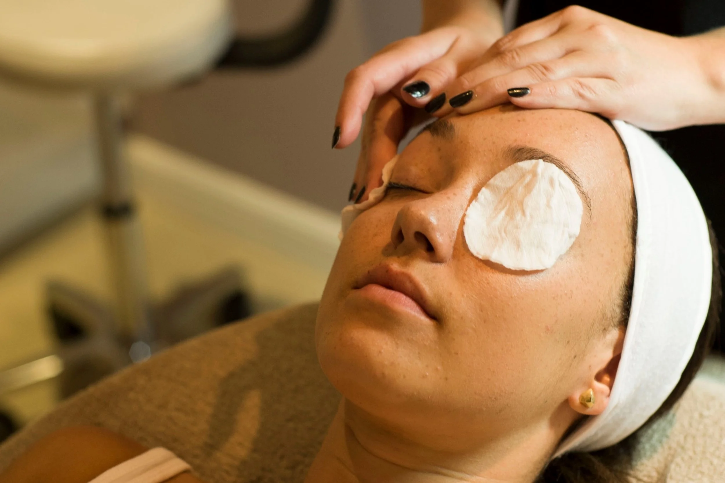 A woman receives a facial treatment, with a cotton pad on her right eye, while lying down with her head supported and her eyes closed.