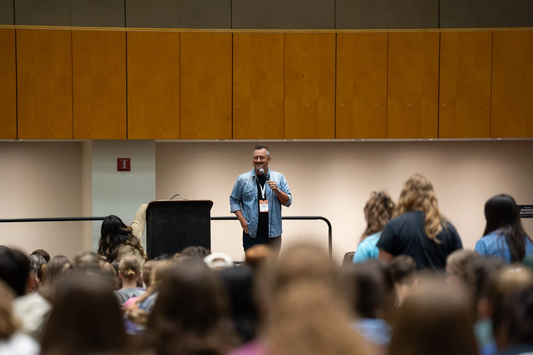 A man standing on a stage holding a microphone, smiling, in front of a large audience in a conference room.