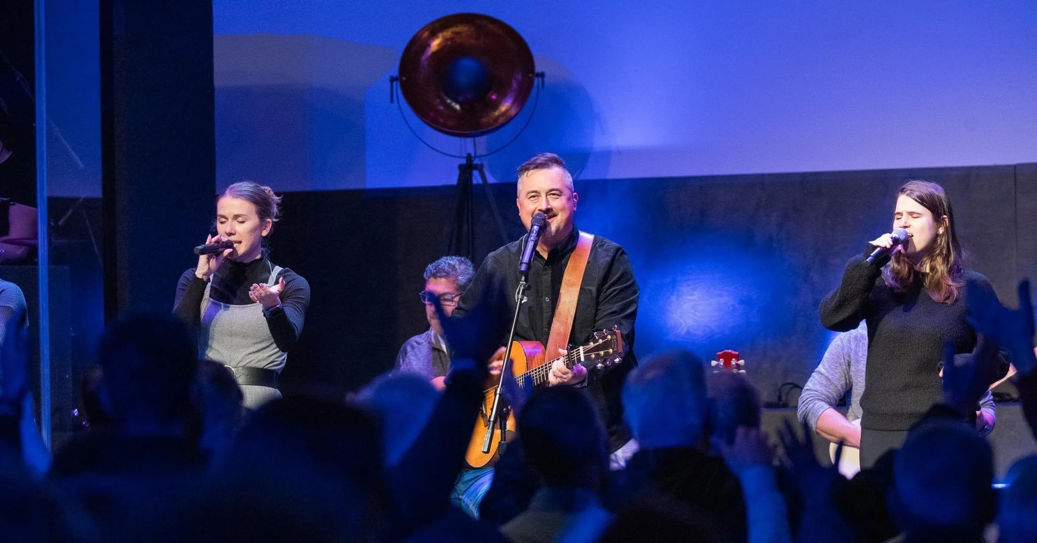 Musicians performing on stage with an audience in the foreground, a man playing guitar and singing, flanked by two women singing into microphones, with a large lighting fixture behind them.