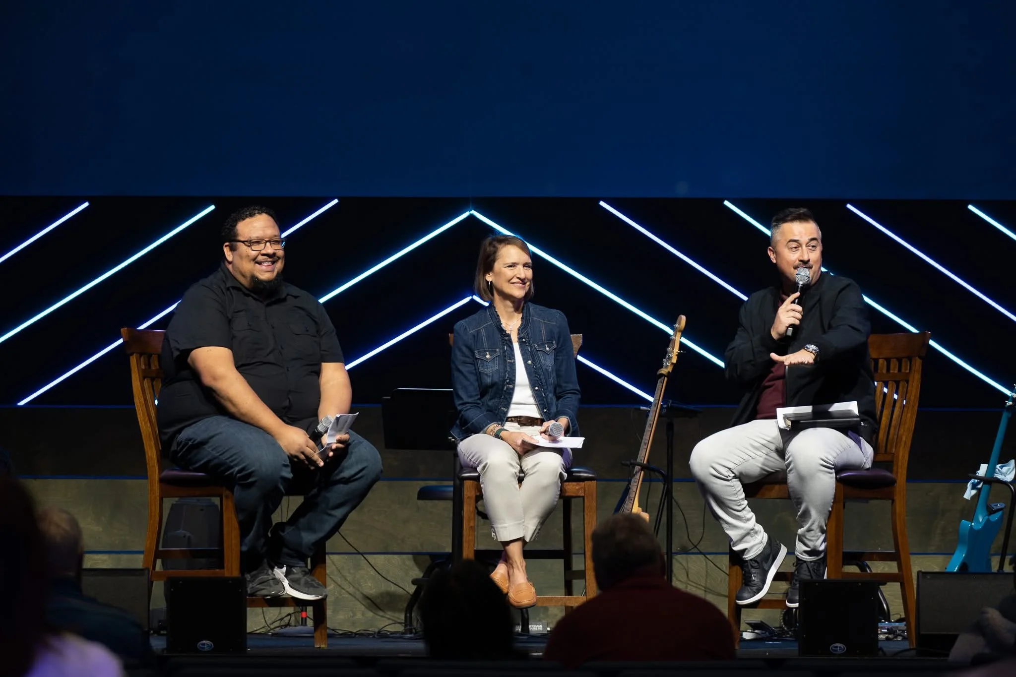 Three people sitting on a stage with a patterned background, engaging in a panel discussion, with microphones in hand.