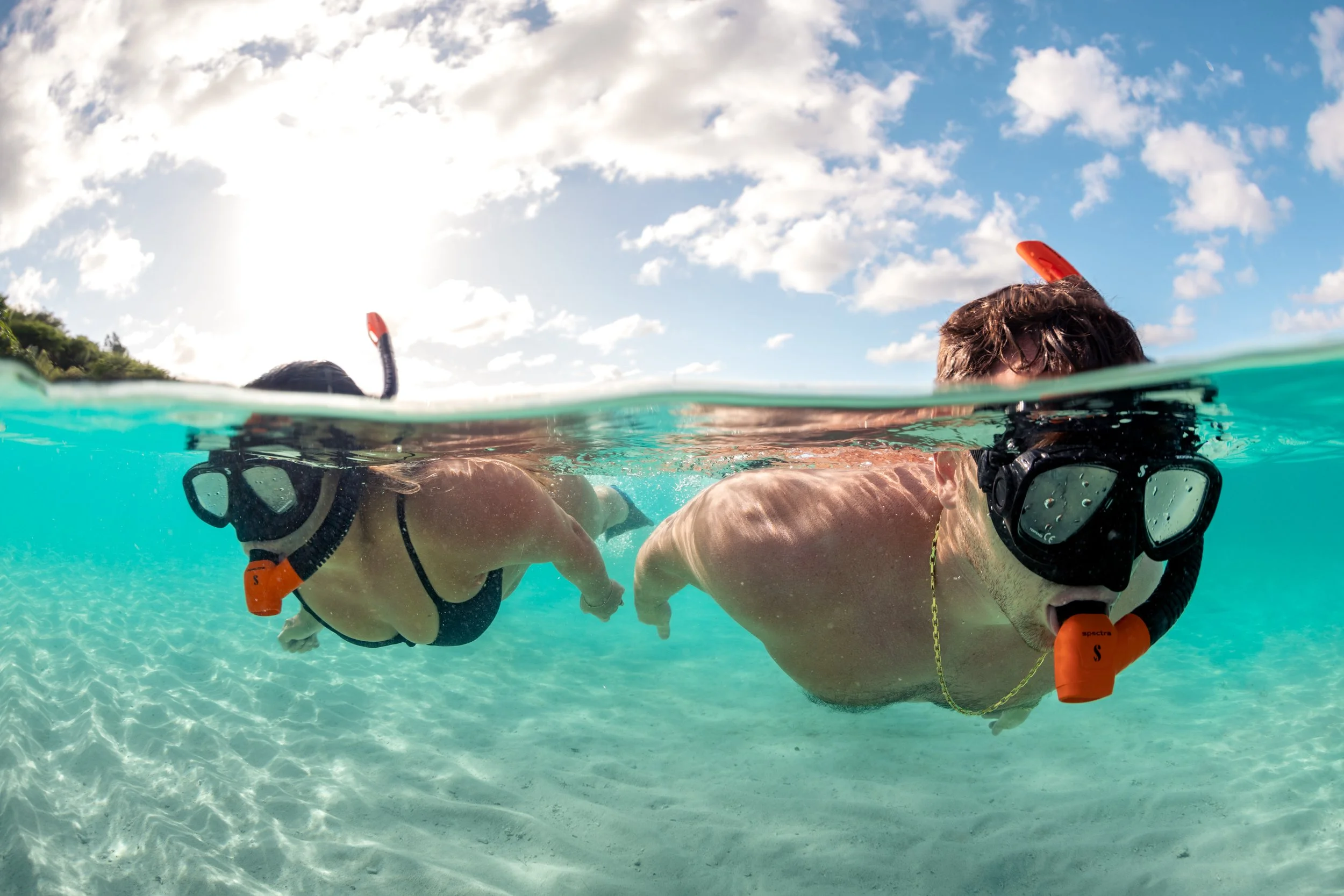 Two people snorkeling in clear turquoise water, wearing masks and snorkels, with a partly cloudy sky overhead.