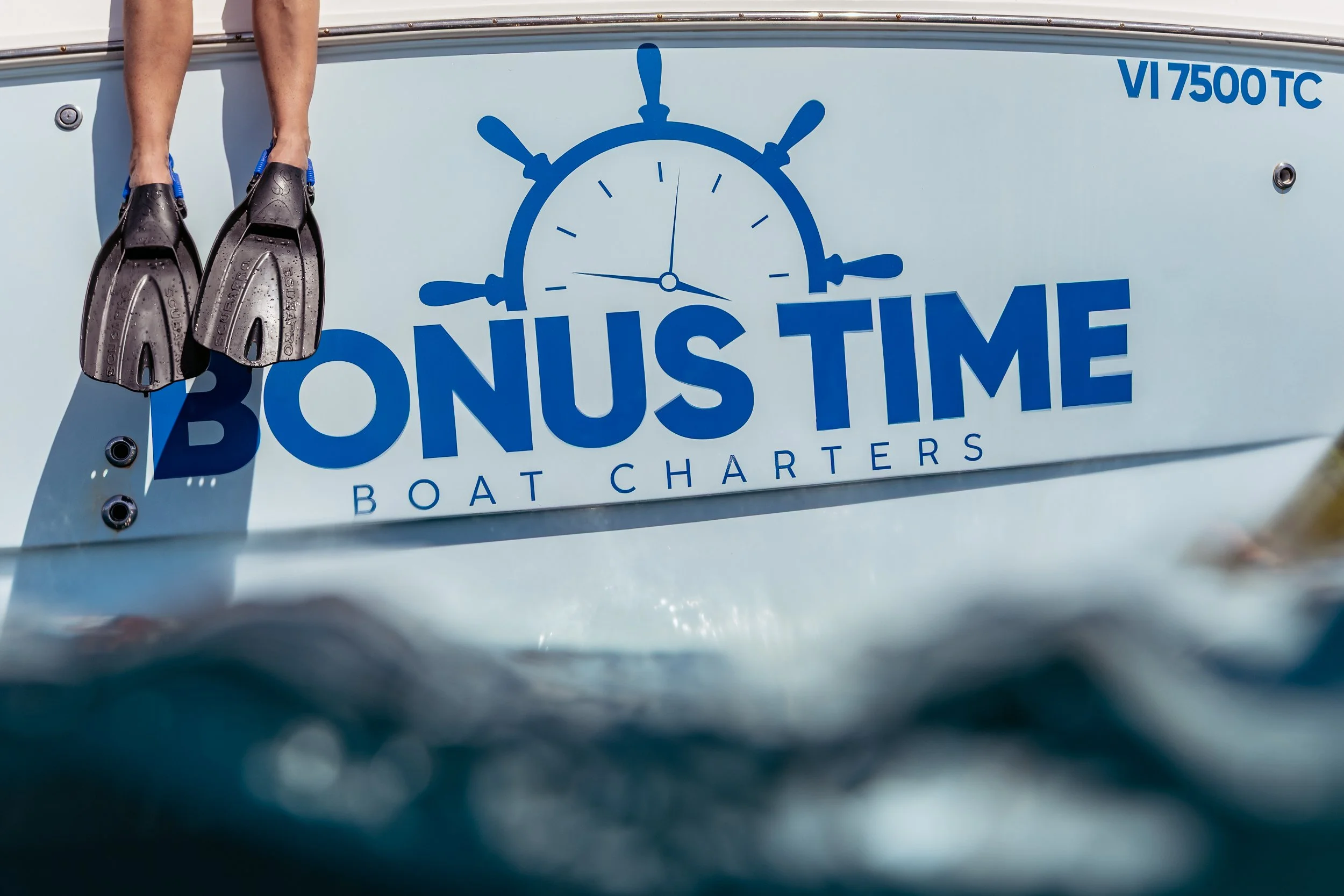 Close-up of a boat with the words "Bonus Time Boat Charters" and a clock logo on the side, with a pair of swim fins hanging over the edge of the boat, taken from the water.