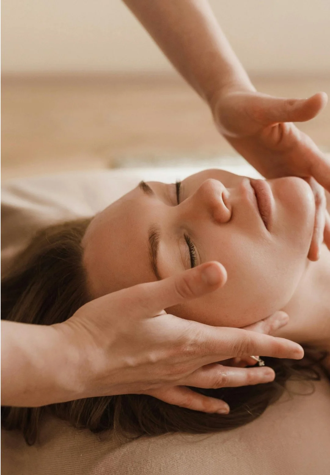 A woman with closed eyes receiving a facial massage from a therapist, lying on a massage table.
