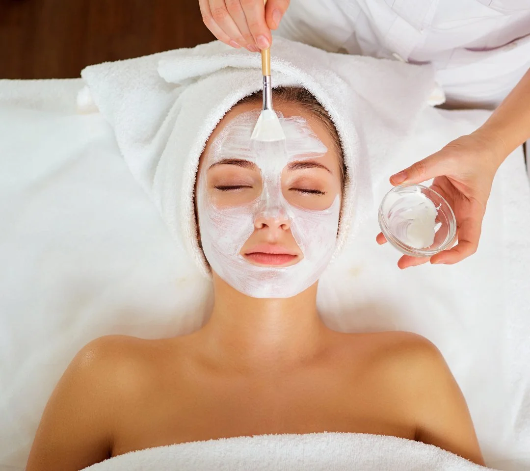 A woman is lying down with a towel wrapped around her head, receiving a facial treatment. A skincare professional is applying a white facial mask to her face with a spatula, holding a bowl of the mask in their other hand.
