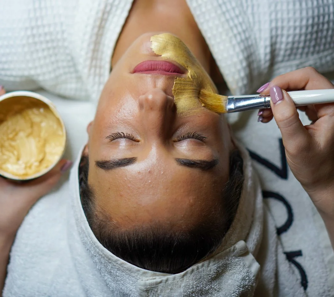 A woman receives a facial treatment with a gold-colored mask applied to her face, lying down with closed eyes, with a spa towel wrapped around her head.
