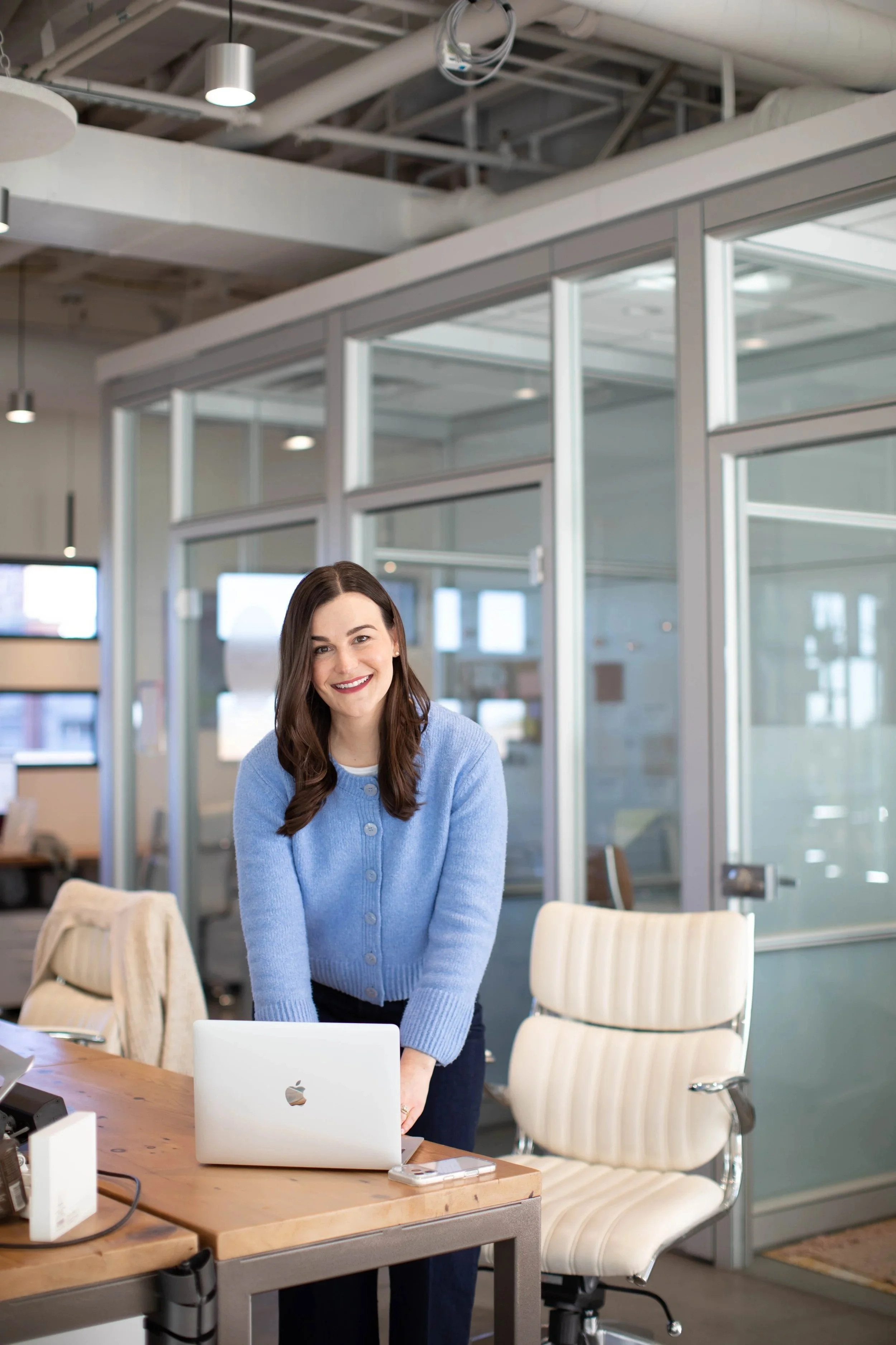 Smiling woman with long dark hair wearing a blue sweater leaning over laptop in an office background