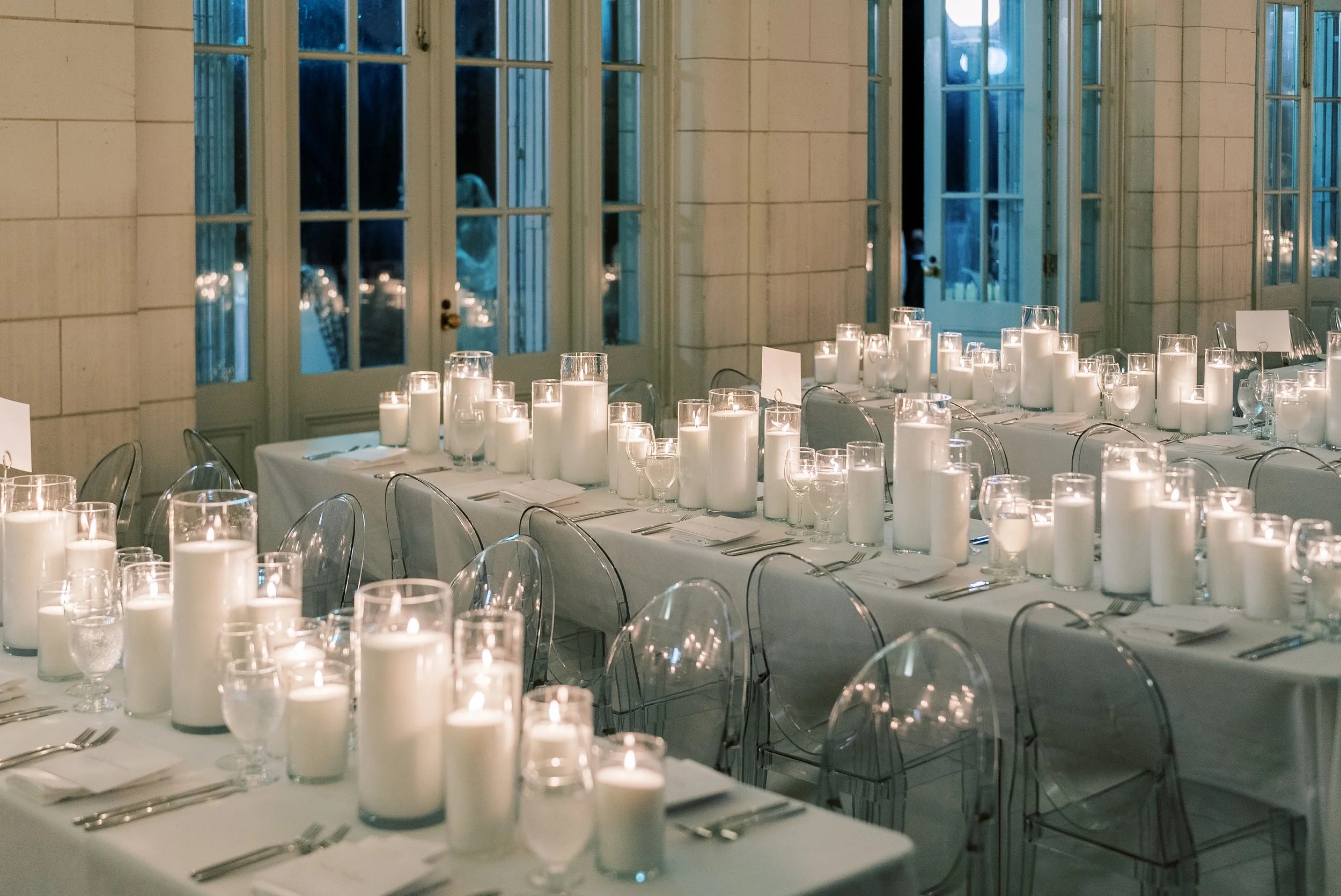 Elegant event setup with long tables covered in white tablecloths, decorated with numerous pillar candles and glassware, illuminated by soft lighting, inside a room with large glass doors and windows at night.