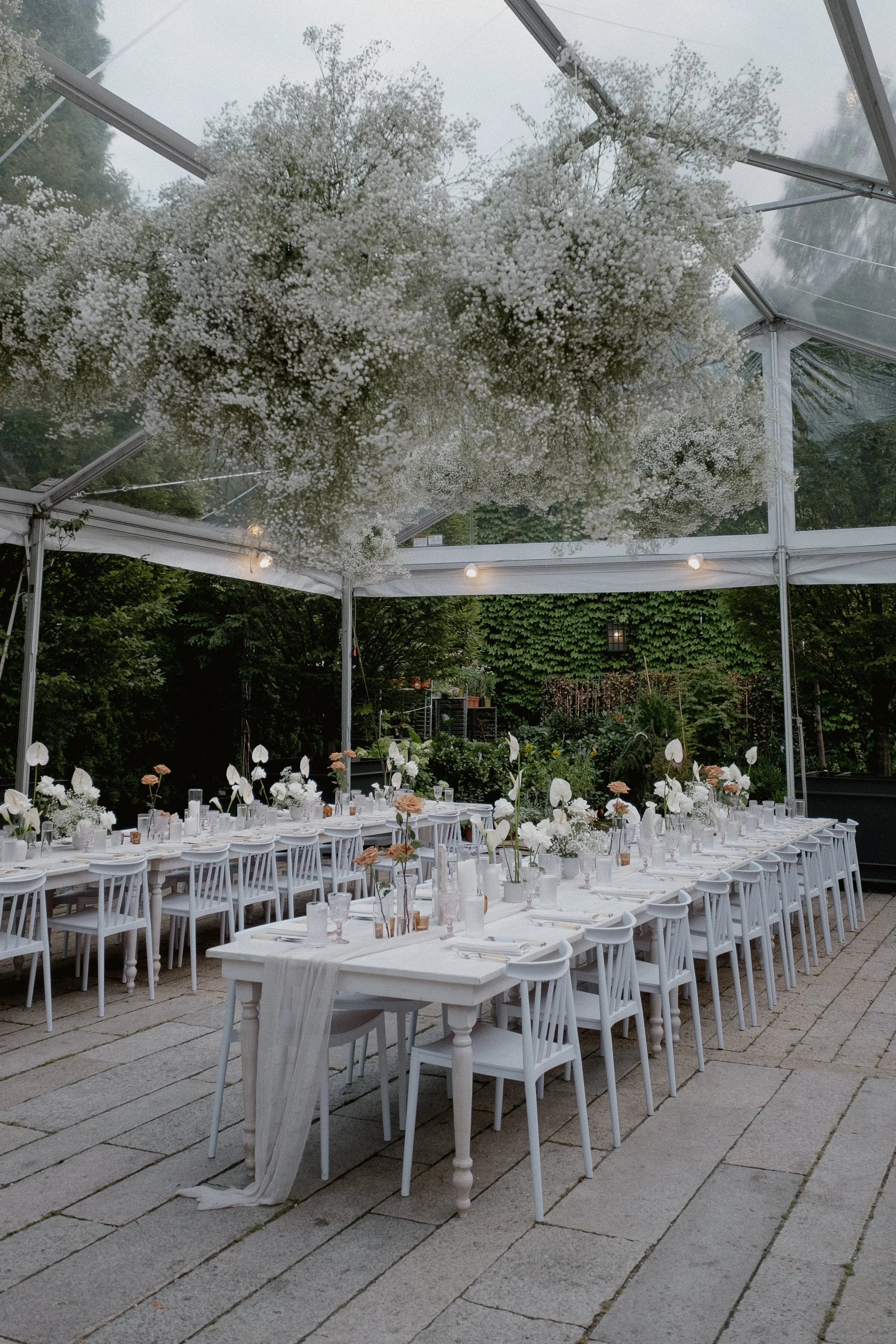 Empty outdoor wedding reception area under a transparent tent with white tables, chairs, and floral centerpieces, surrounded by greenery.