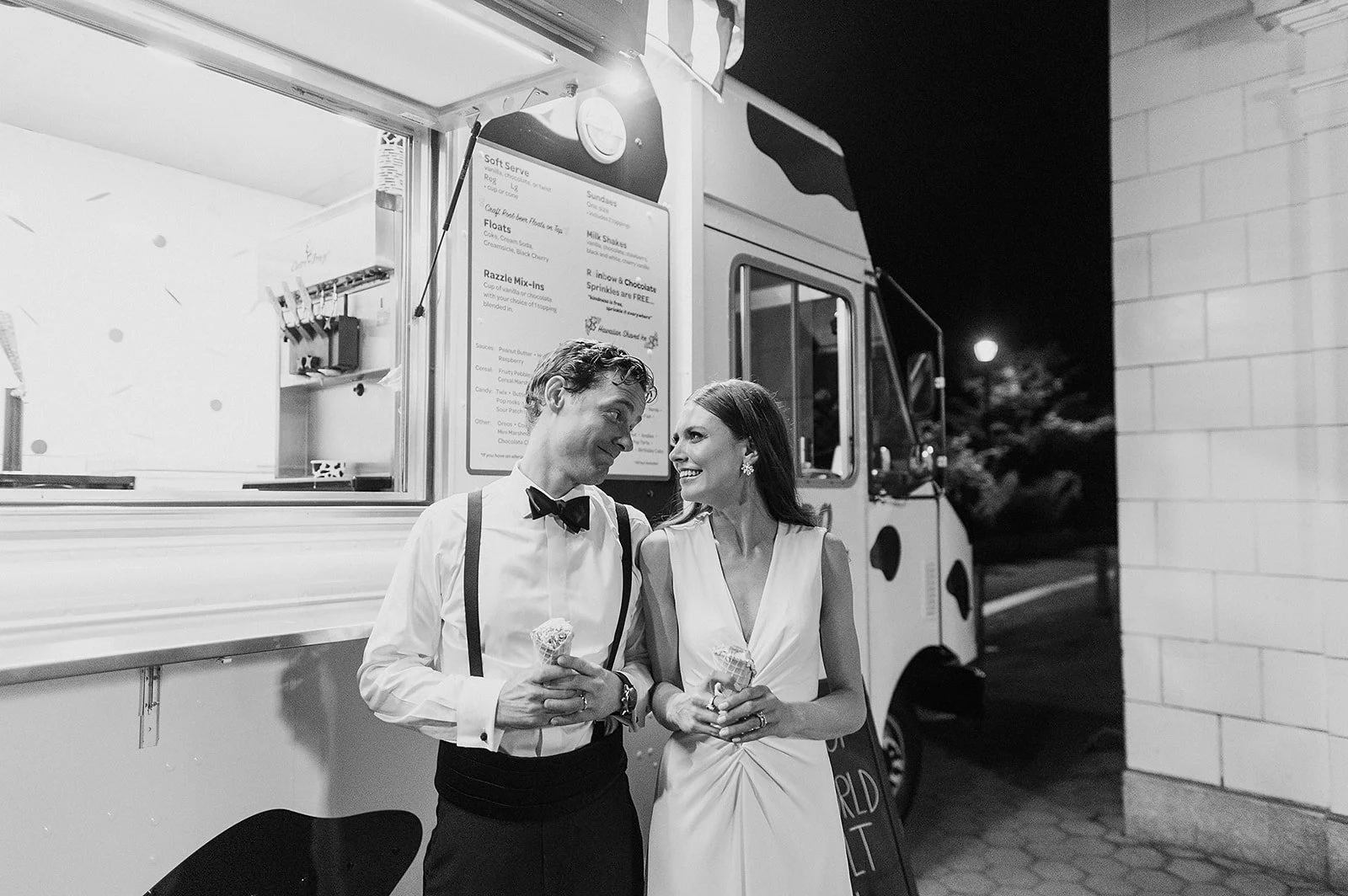 A black and white photo of a man and woman in formal attire standing outside a dessert truck at night, smiling at each other and holding ice cream cones.