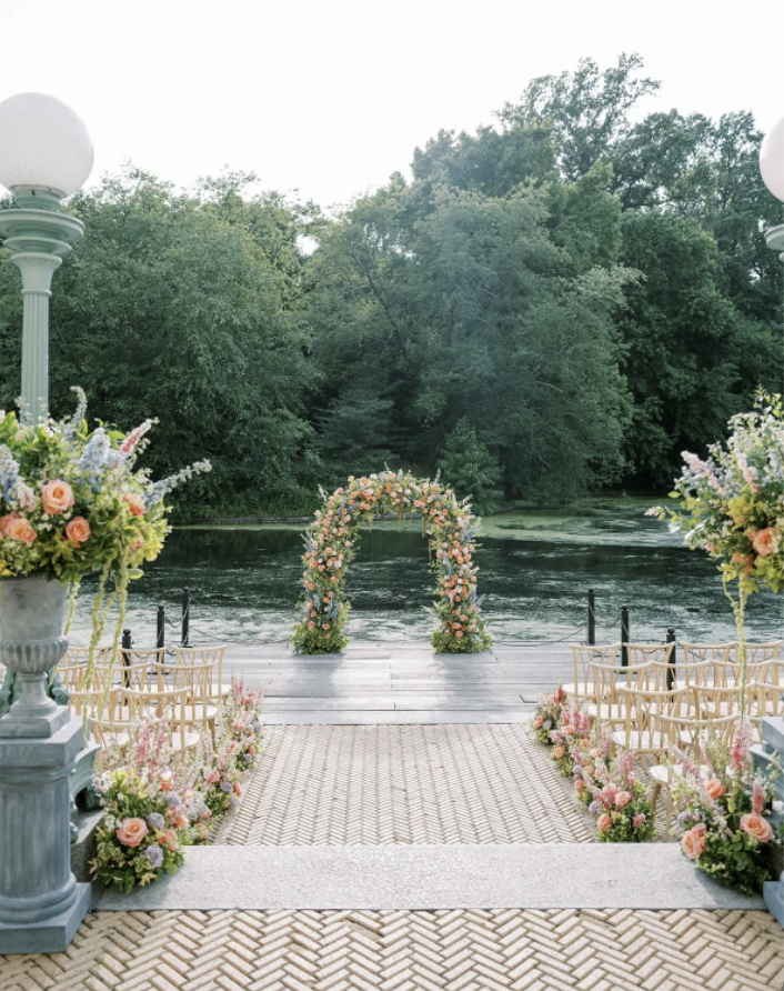 Wedding ceremony setup on a brick pathway by a river with an arch decorated with flowers, floral arrangements on pedestals, and outdoor chairs on either side.