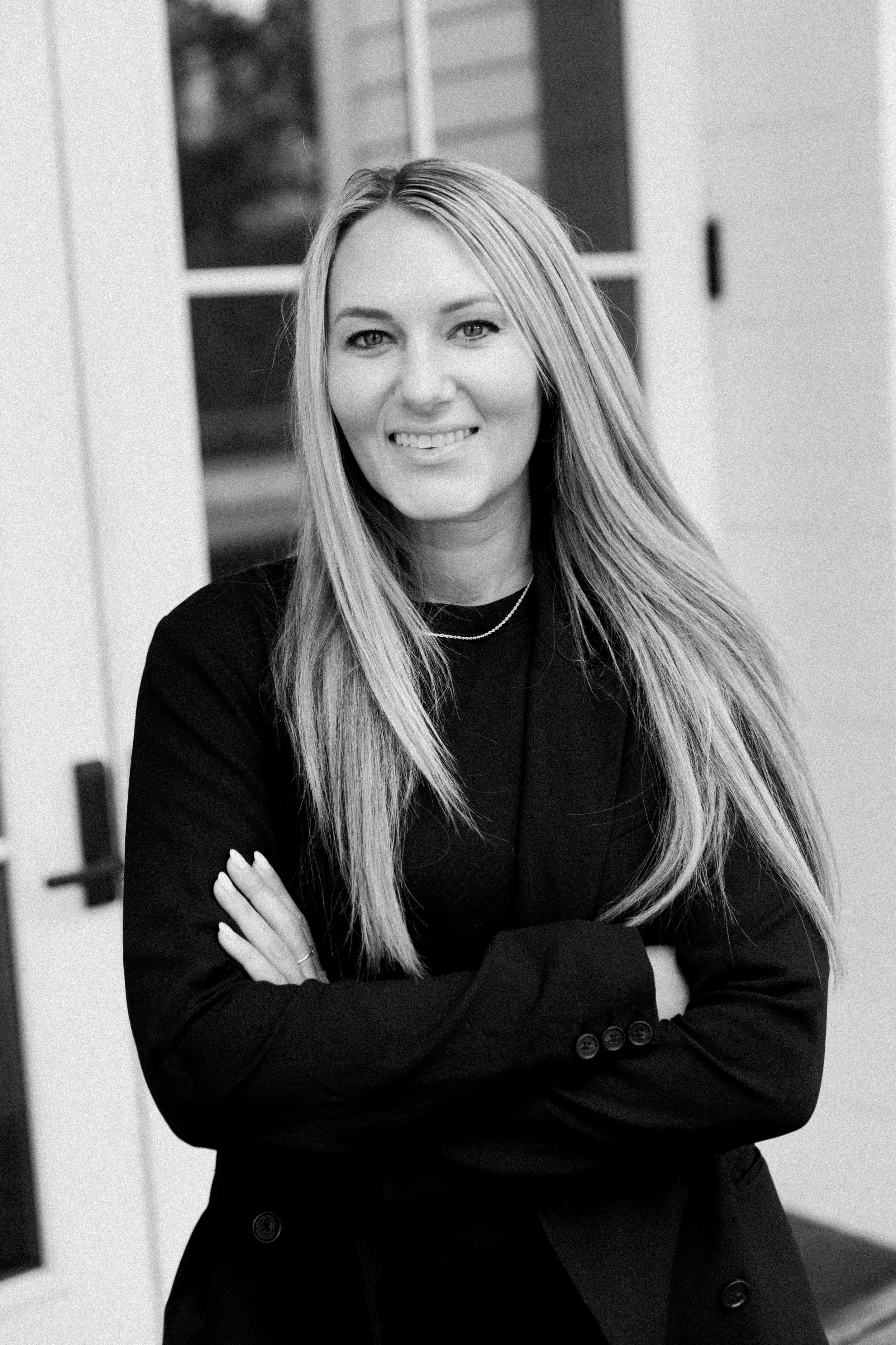 A black-and-white photo of a woman with long blonde hair, smiling, wearing a black blazer and crossed arms, standing in front of a window and door.