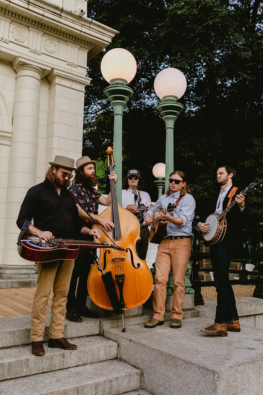 A group of five musicians playing string and folk instruments outdoors, standing on steps near a classic building with decorative columns and street lamps, during evening or dusk.