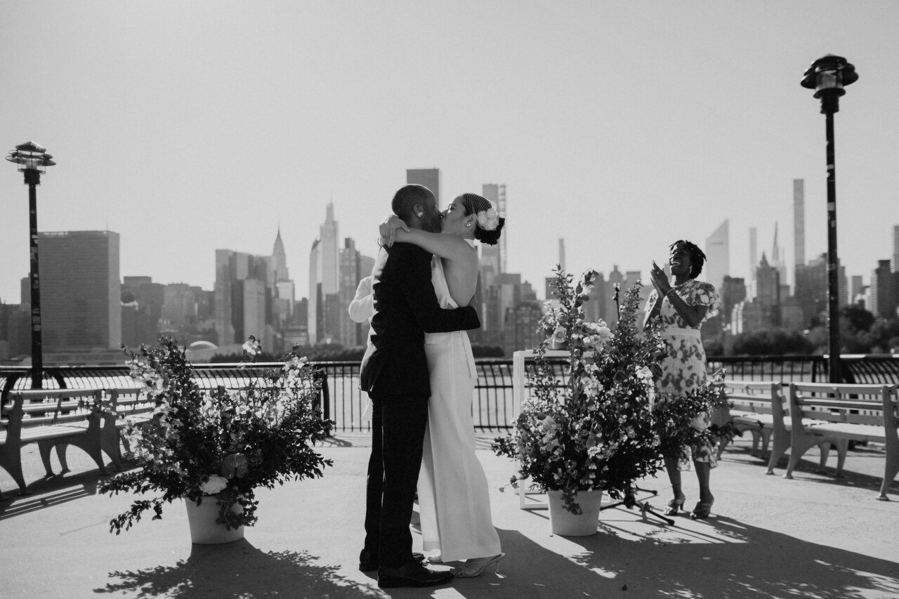 A couple in wedding attire sharing a kiss during their outdoor wedding ceremony with a city skyline in the background, a woman standing nearby clapping and smiling.
