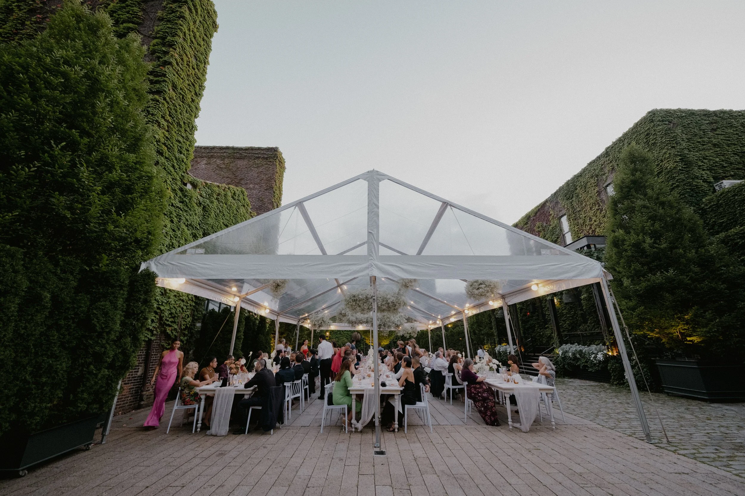 Outdoor wedding reception under a white tent with guests seated at long tables, surrounded by green ivy-covered walls and buildings, evening setting with soft lighting.