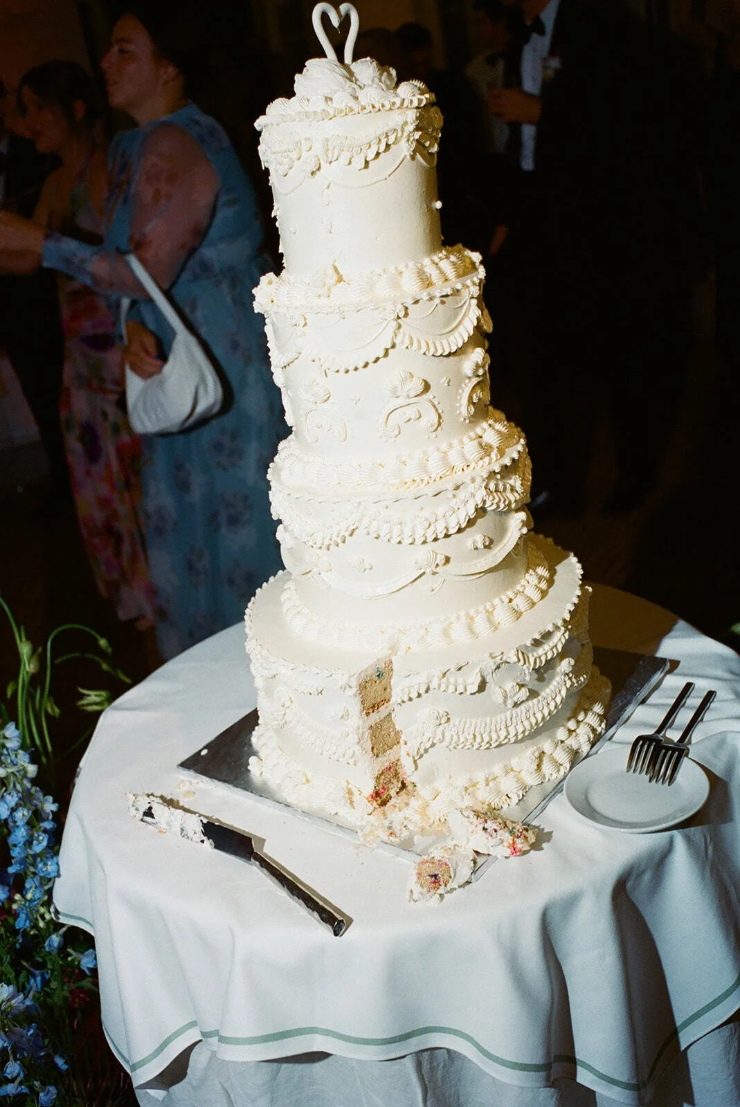 Large, multi-tiered white wedding cake with intricate piping and decorative details, placed on a white tablecloth-covered table with a cake knife and empty plates nearby; a piece has been cut from the cake, revealing layers inside.