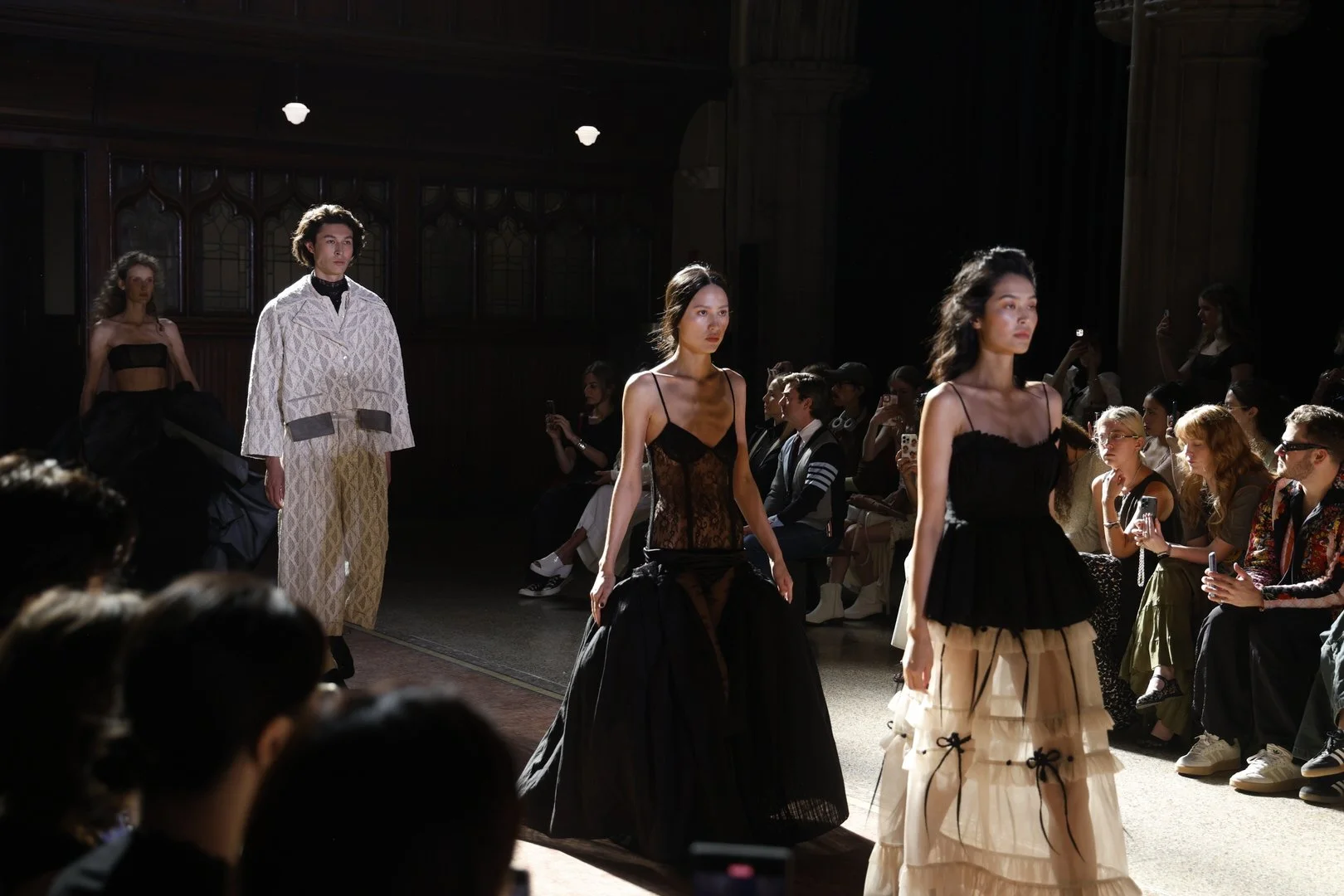 Models walking down runway during a fashion show, audience seated on either side watching