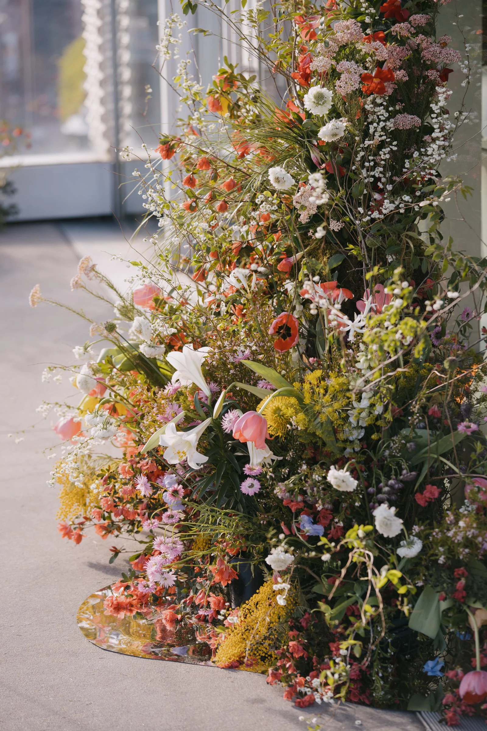 A large, colorful bouquet of various flowers arranged on the ground near glass doors, with sunlight illuminating the scene.
