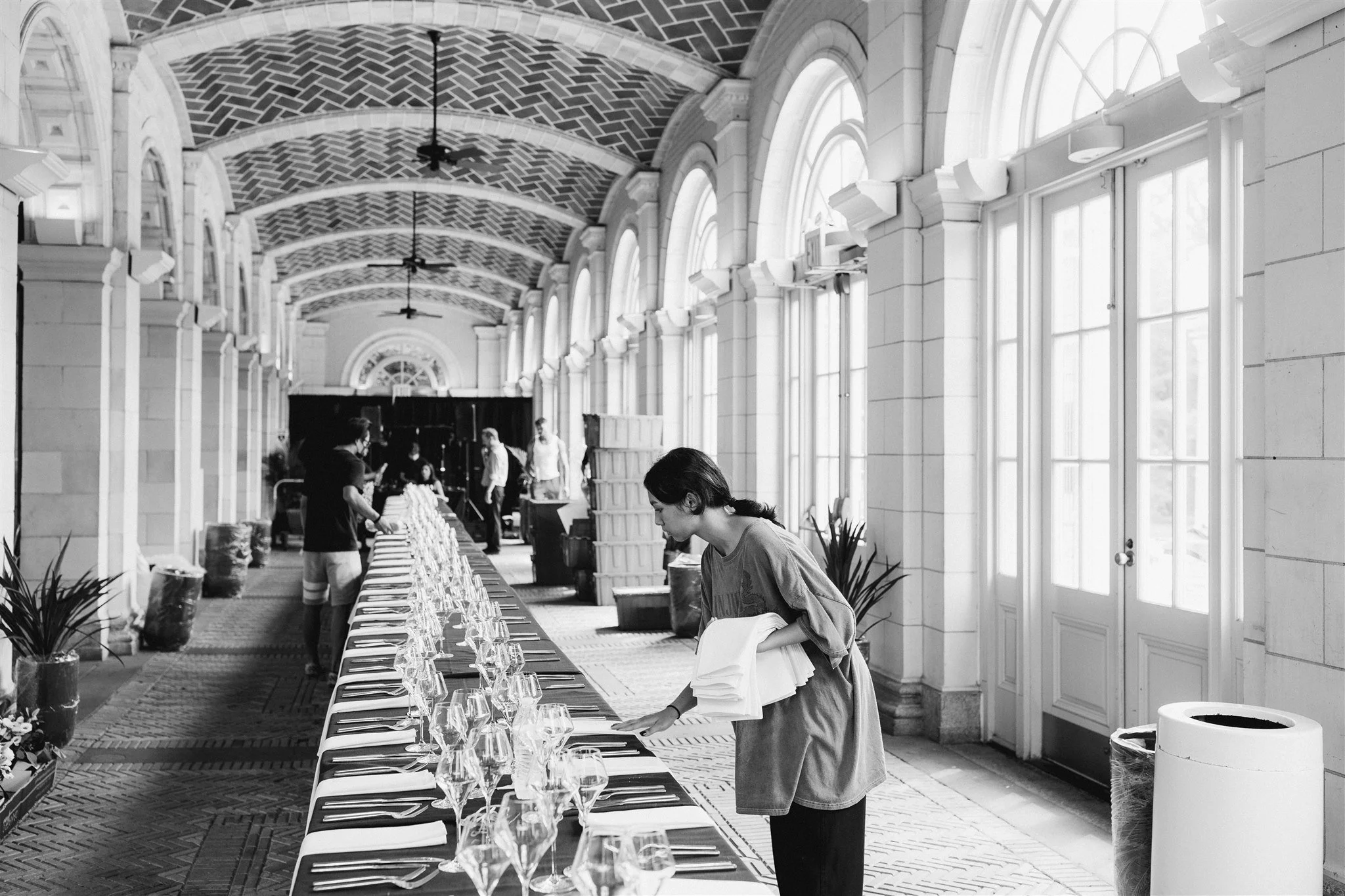 Black and white photo of a long banquet table set with glasses and cutlery, inside a bright hall with large arched windows, where a woman is tidying the table while others prepare in the background.