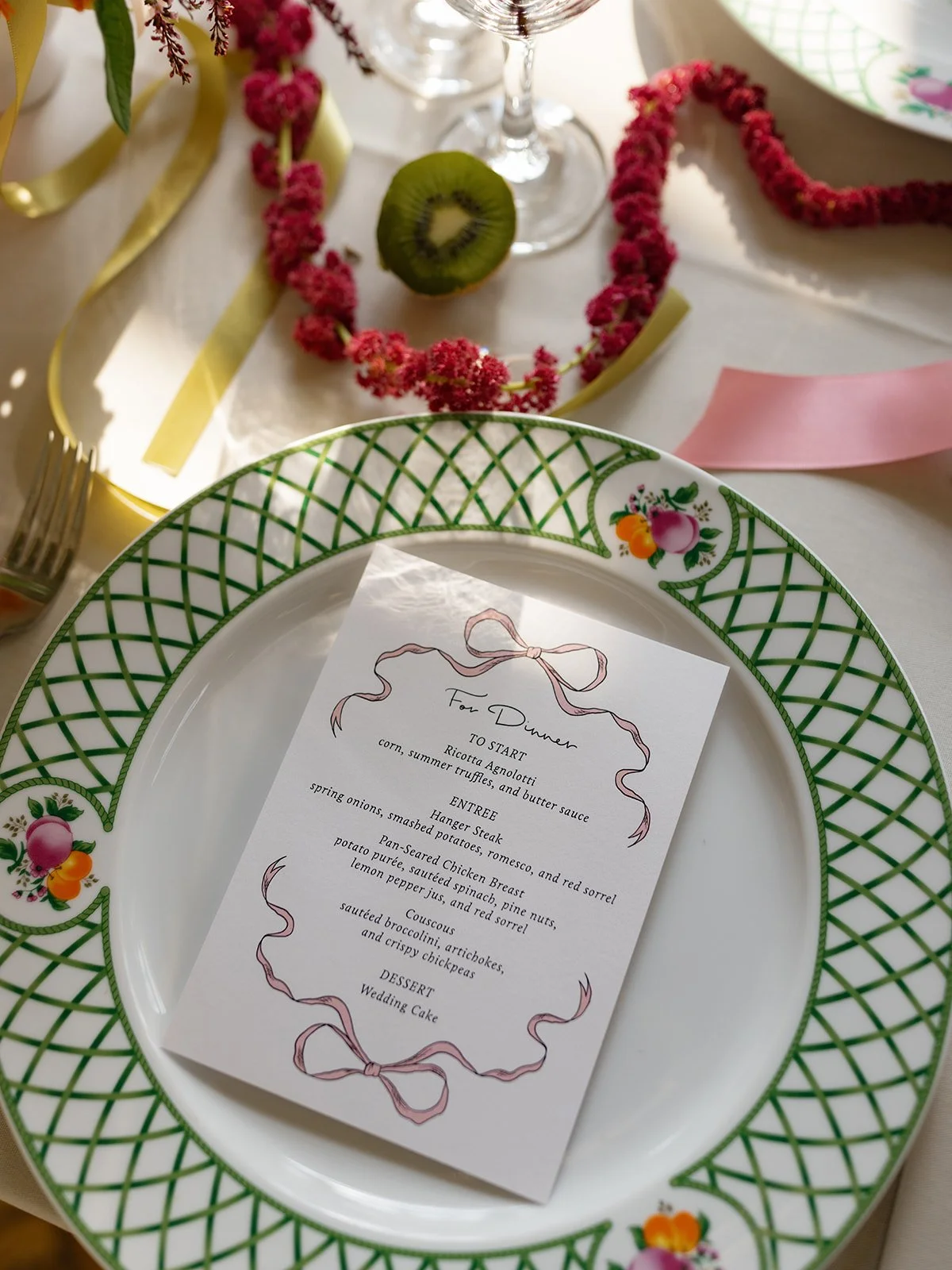 A dinner table setting with a white plate bordered with green lattice pattern and floral designs. A printed menu is placed on the plate, and the table is decorated with pink flowers, a halved kiwi, a wine glass, yellow ribbon, pink paper, and a pink 