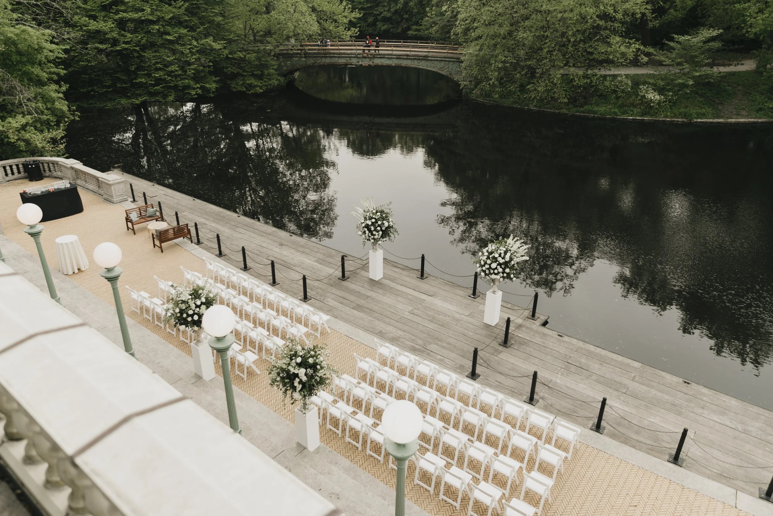 A wedding ceremony setup near a river with white chairs arranged in rows, floral arrangements on white pillars, and black and wooden benches, all on a wooden deck looking over water and surrounded by green trees.
