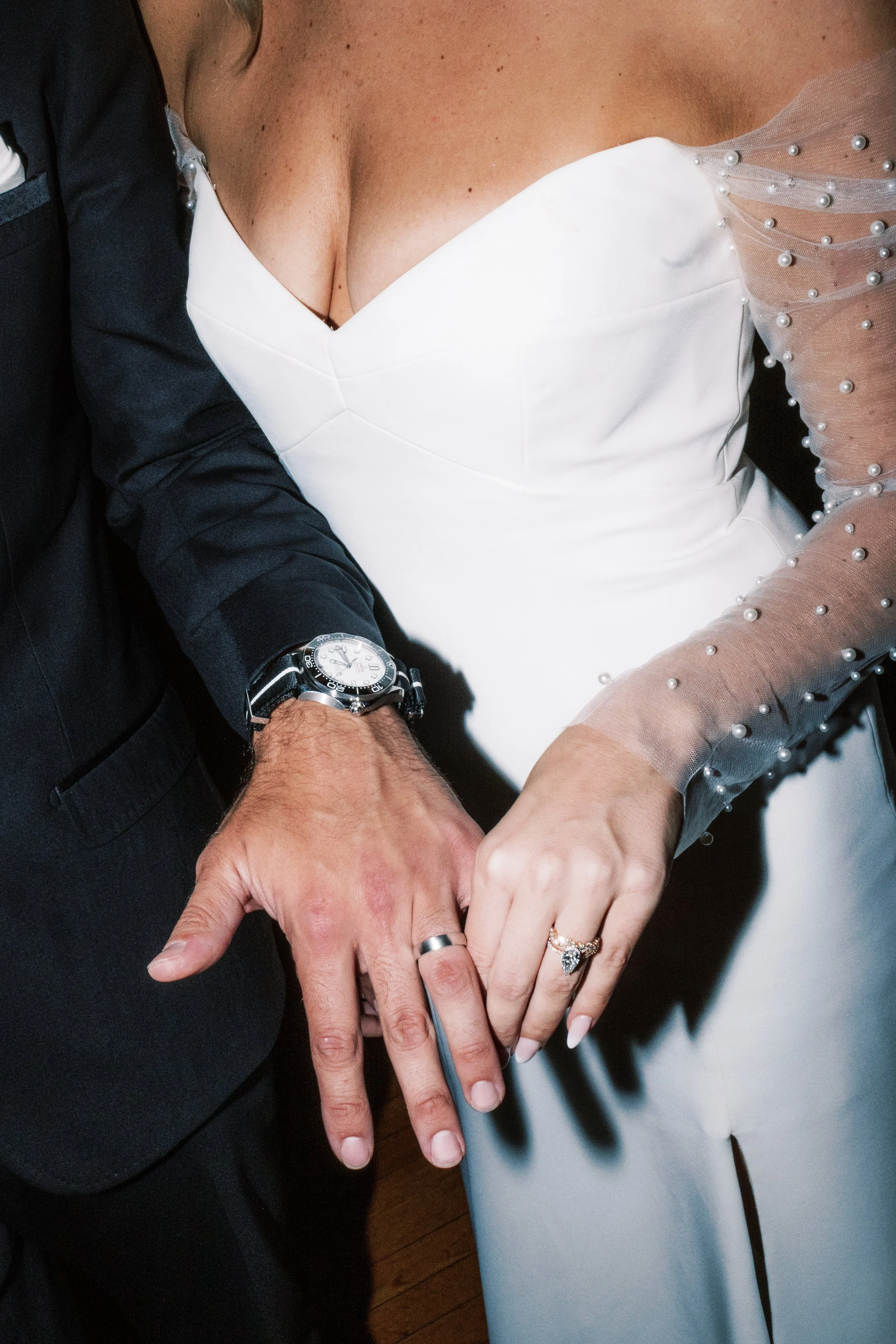 Close-up of a couple holding hands, with wedding rings visible. The woman is wearing a white wedding gown with pearl embellishments on the sheer long sleeve and a ring with a large stone. The man's hand shows a wedding band and an elaborate watch.