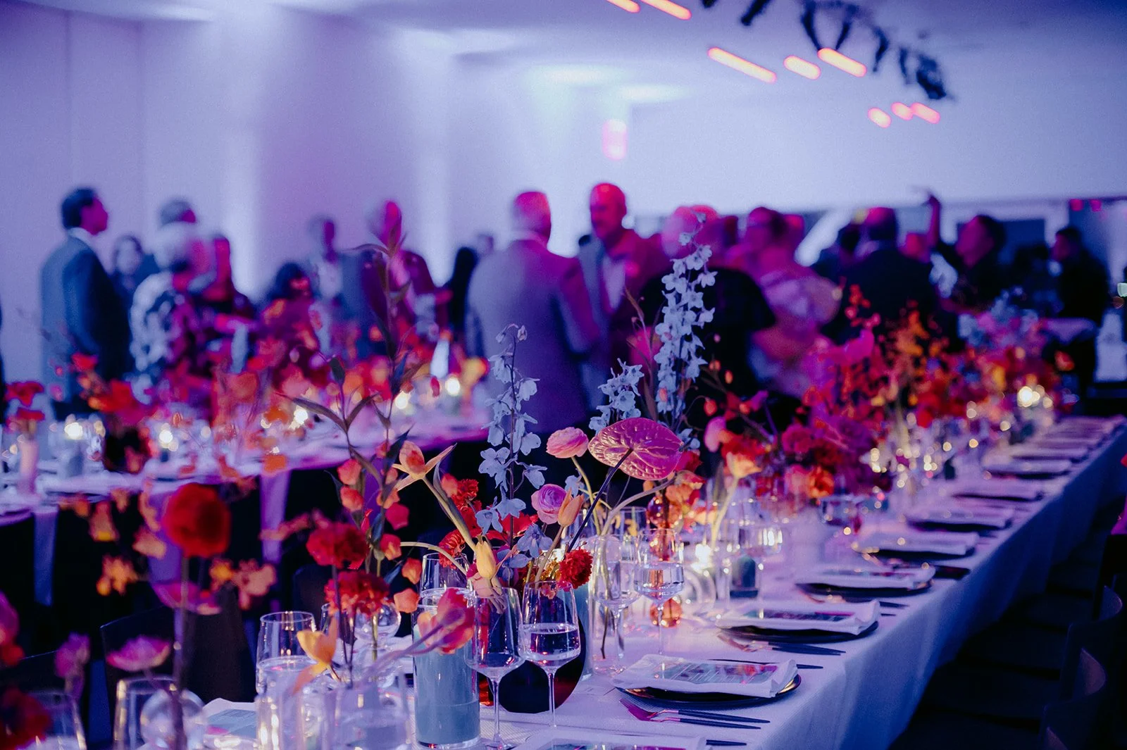 A long banquet table adorned with colorful floral arrangements and wine glasses at a formal event or celebration, with guests mingling in the background.