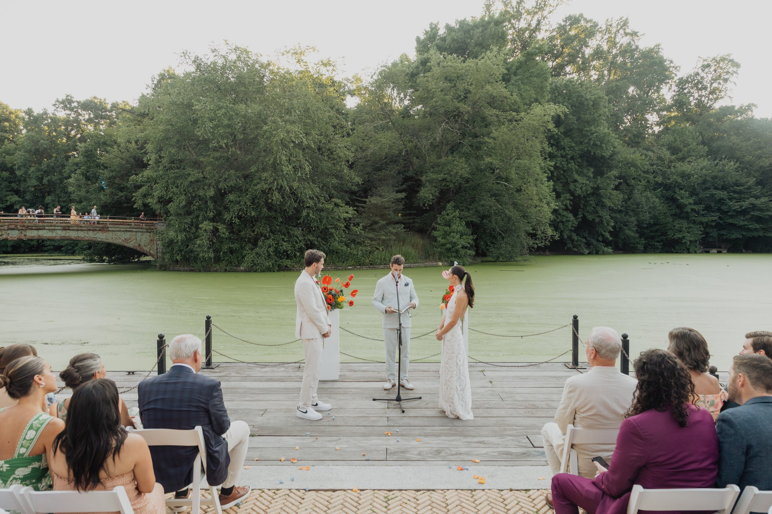 A wedding ceremony taking place outdoors on a wooden dock over a pond surrounded by green trees. The bride and groom stand facing each other with the officiant in the middle under a microphone, holding flowers. Guests are seated on white chairs facin