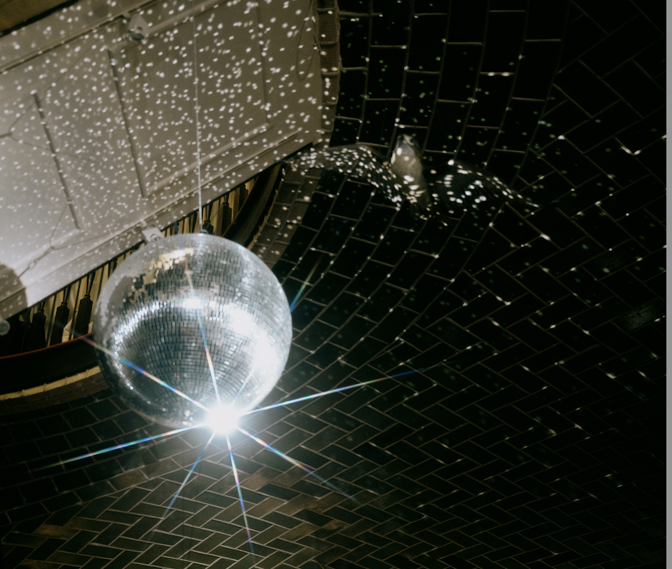 A disco ball hanging from the ceiling with a bright light reflecting off of it, casting small light spots on the dark tiled ceiling.