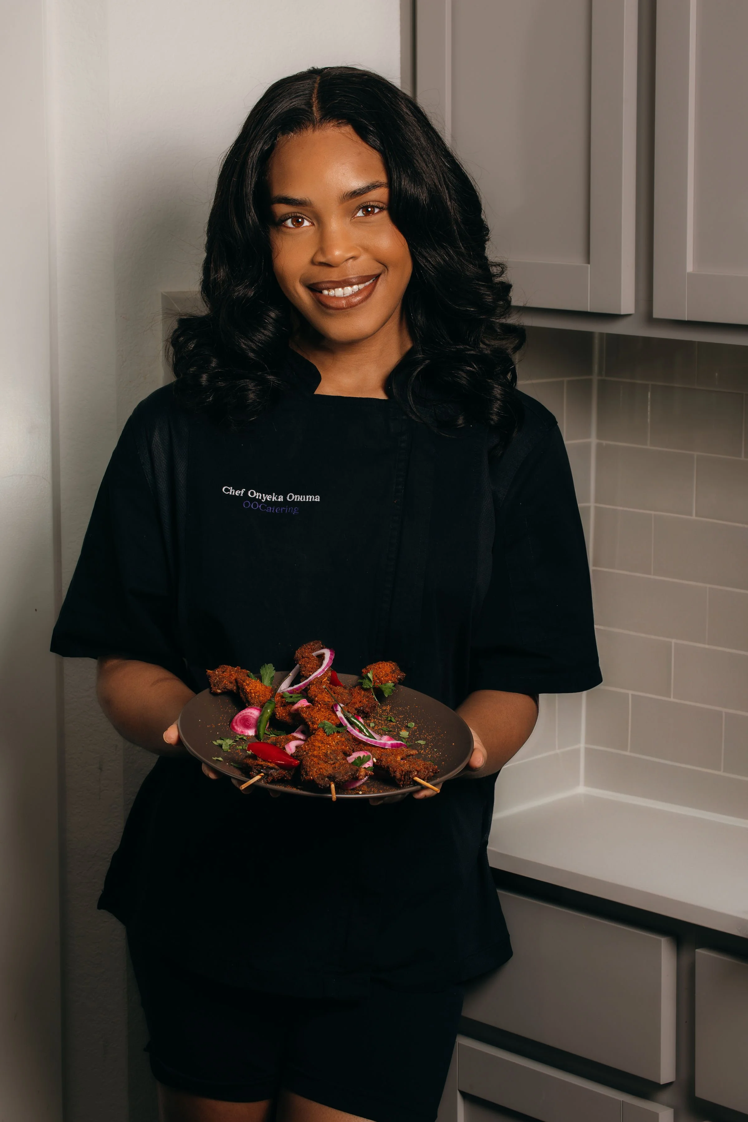 A woman in a black chef uniform holding a plate of skewered fried food garnished with herbs and sliced vegetables, standing in a kitchen.