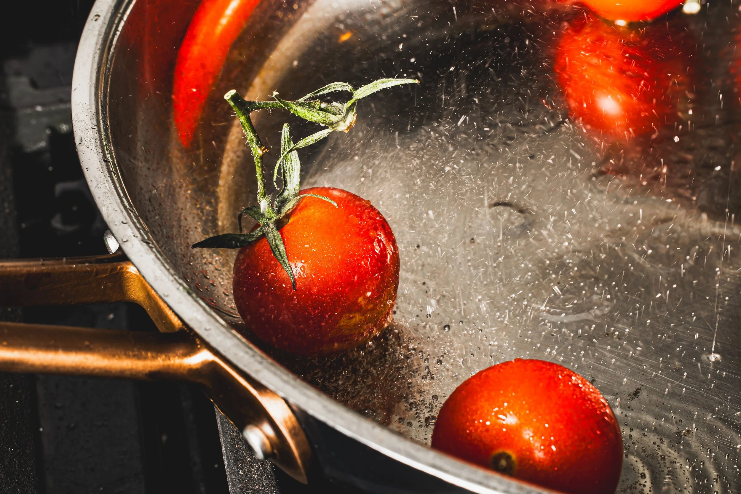 Tomatoes being washed in a stainless steel pan as water splashes around.