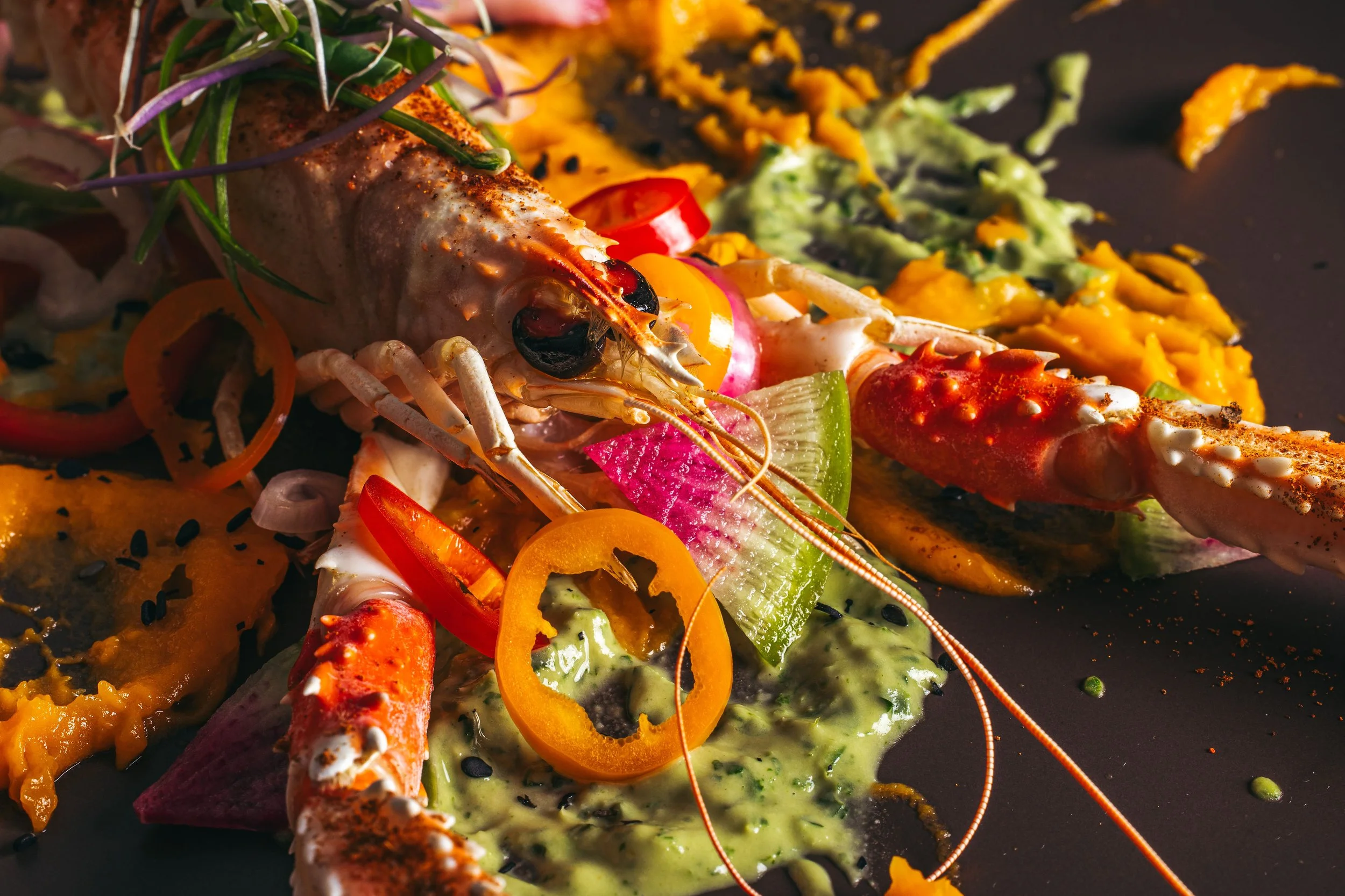 Close-up of a gourmet dish featuring a cooked lobster tail with various colorful vegetables and sauces on a black plate.