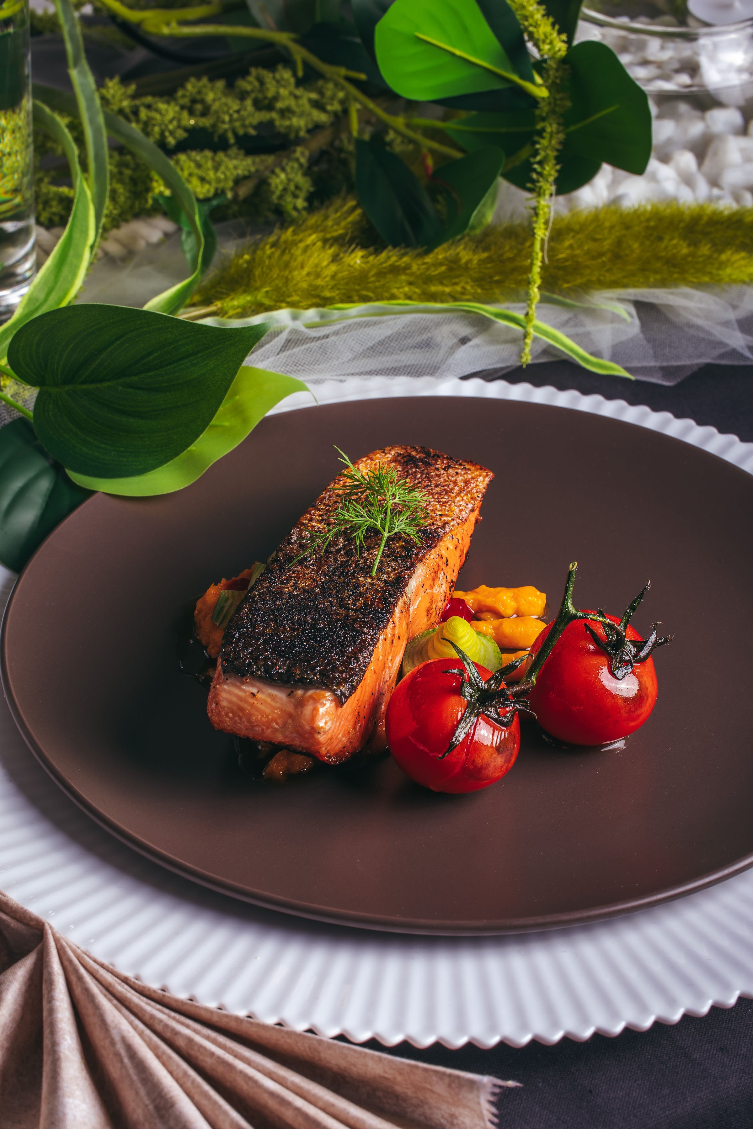 A plated dish featuring a piece of seared salmon with crispy skin, garnished with a sprig of dill, accompanied by grape tomatoes and a vegetable side, set on elegant tableware with green plants in the background.